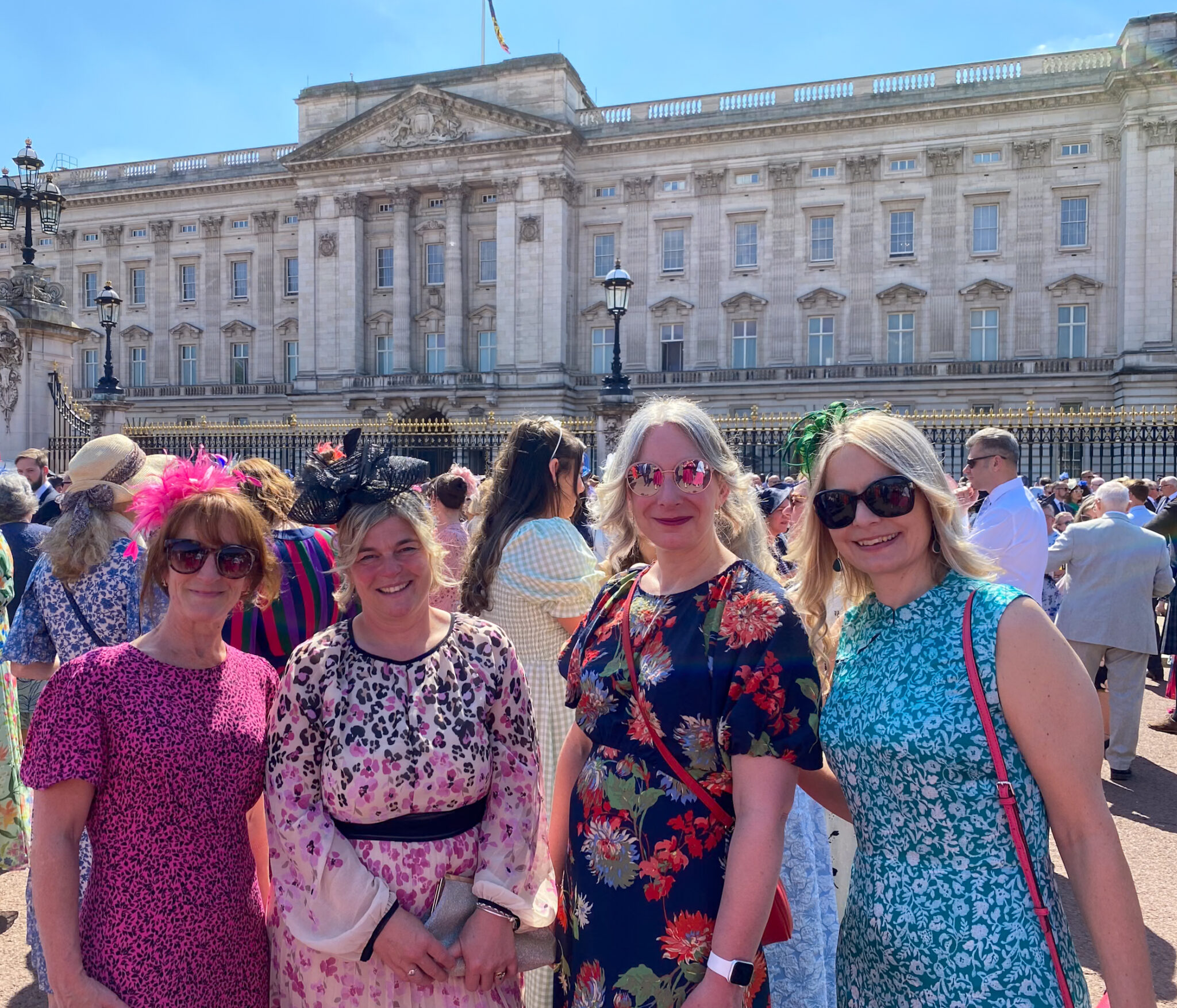 Four people dressed in vibrant, formal summer attire stand smiling in front of Buckingham Palace on a clear, sunny day. Each person is wearing a unique outfit with bright patterns and floral designs, along with decorative fascinators or headpieces. Two - far left and on the right - wear sunglasses and all appear to be enjoying the festive atmosphere, surrounded by a well-dressed crowd gathered outside the ornate palace gates.