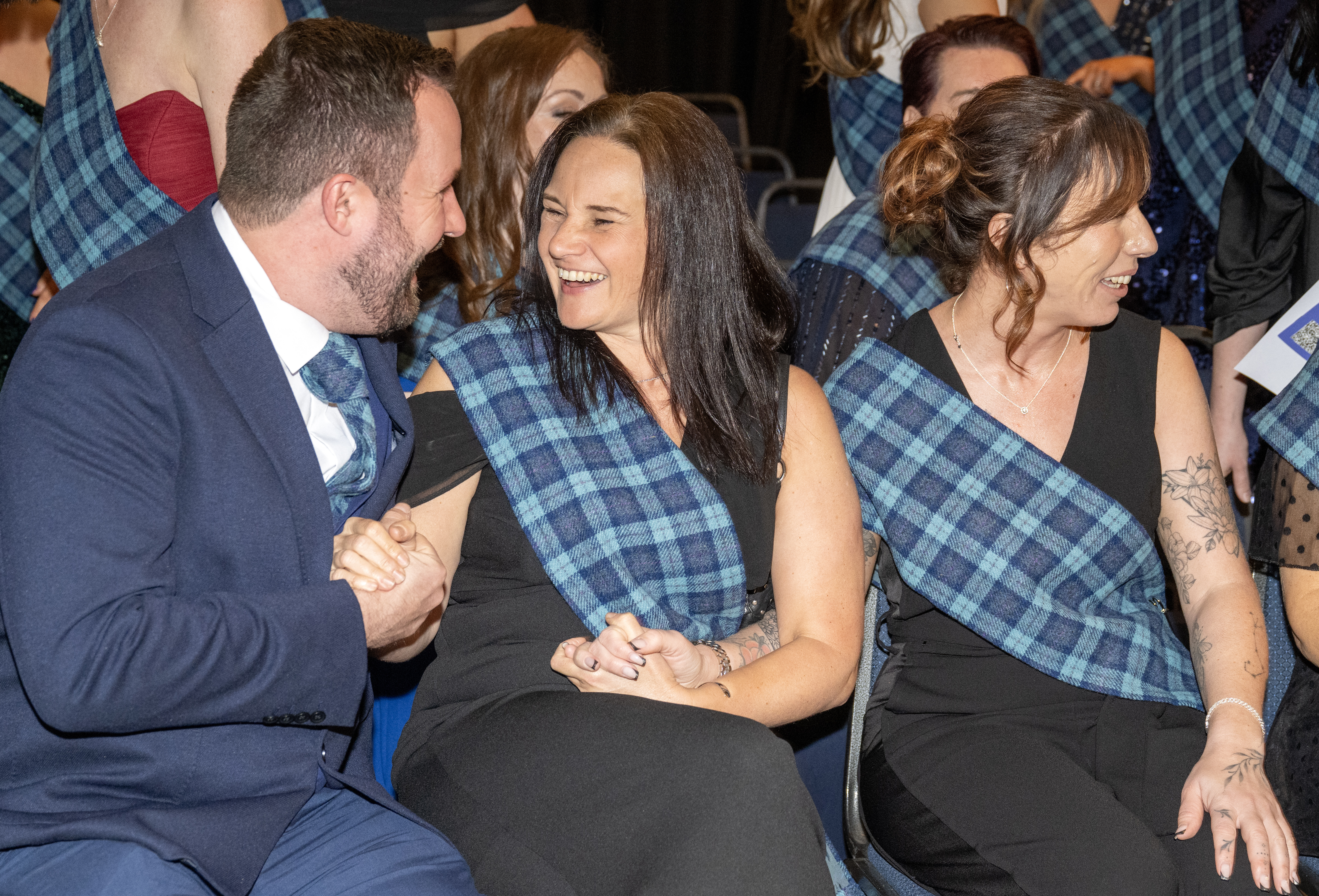 A group of people dressed in formal attire sit closely together, smiling and laughing. Most are wearing black with blue tartan sashes across their shoulders. The person on the left is in a suit and shaking hands warmly with the person next to them, who is wearing a black outfit and smiling broadly. The third person is also dressed in black and looking toward the others with a smile. The background shows more people in similar attire, suggesting a celebratory or formal group event.