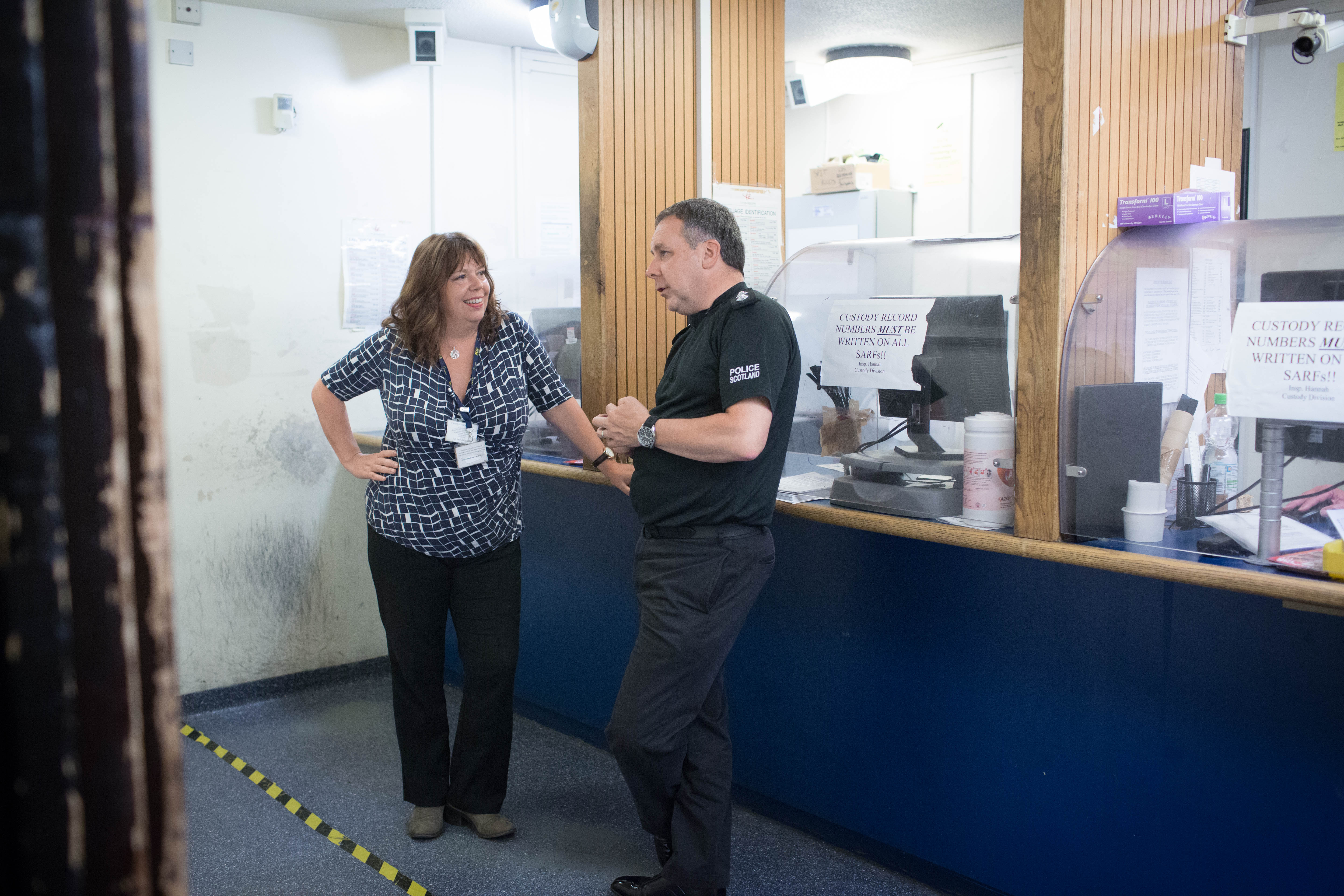 Two people are standing having a conversation in what appears to be a police station or custody reception area. One person is wearing a patterned shirt and dark trousers, while the other is dressed in a dark-coloured uniform with 'Police Scotland' written on the sleeve. They are standing near a counter with clear partitions and signs that read 'Custody record numbers must be written on all SARFs!!' The atmosphere appears informal and friendly.