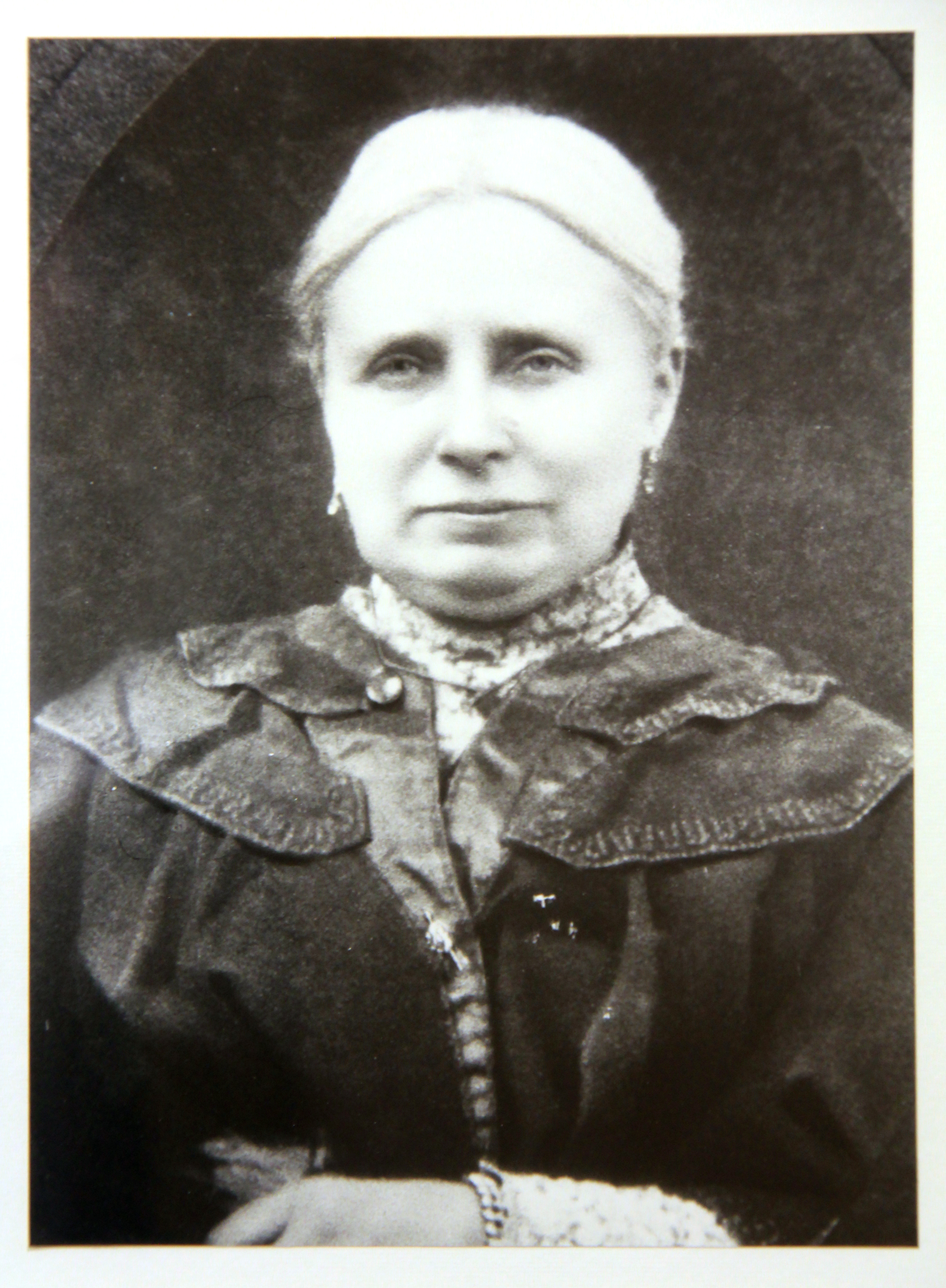 A black-and-white portrait of an older person with light-coloured hair pulled back. They are wearing a dark, high-collared garment with decorative stitching and a lighter patterned undershirt. The person is looking directly at the camera with a neutral expression and has their hands folded in front.