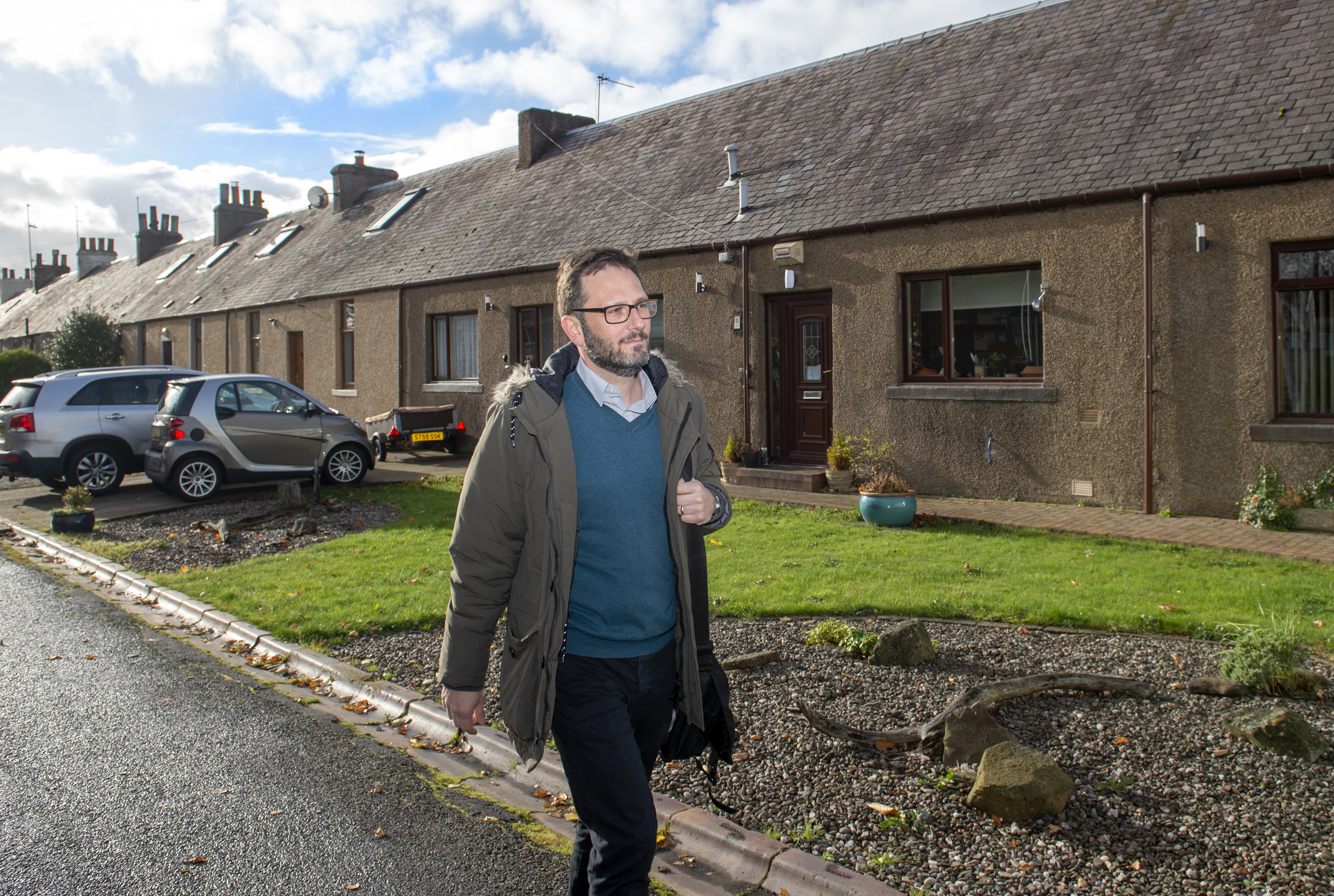 A person with short hair and glasses walks along a residential street on a sunny day. They are wearing a green parka with a fur-lined hood, a teal sweater over a collared shirt, and dark trousers. They carry a bag on one shoulder. Behind them is a row of brown brick terraced houses with sloped roofs and chimneys. Two cars are parked in front of the houses, and the front gardens have grass, gravel, and some planters. The sky is partly cloudy.