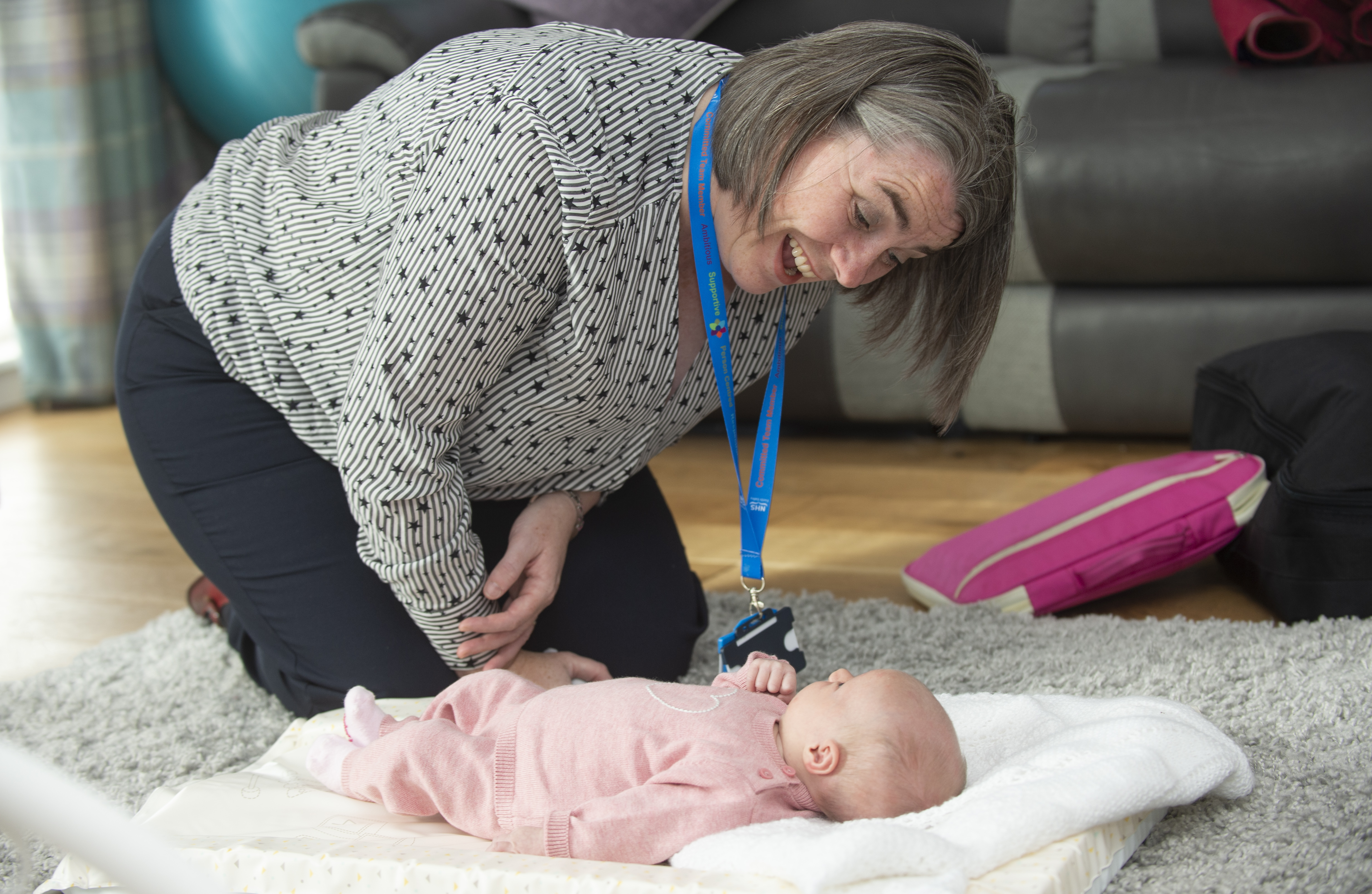 A person with dark grey short hair and wearing a black and white striped blouse with a black star pattern, navy trousers and a blue lanyard kneels on a grey fluffy rug smiling down at a baby who is lying on a white mat that’s on top of the rug. The baby wears a pink babygrow. In the background wooden flooring, a bright pink bag, grey couch and turquoise and lilac plaid curtains are visible.