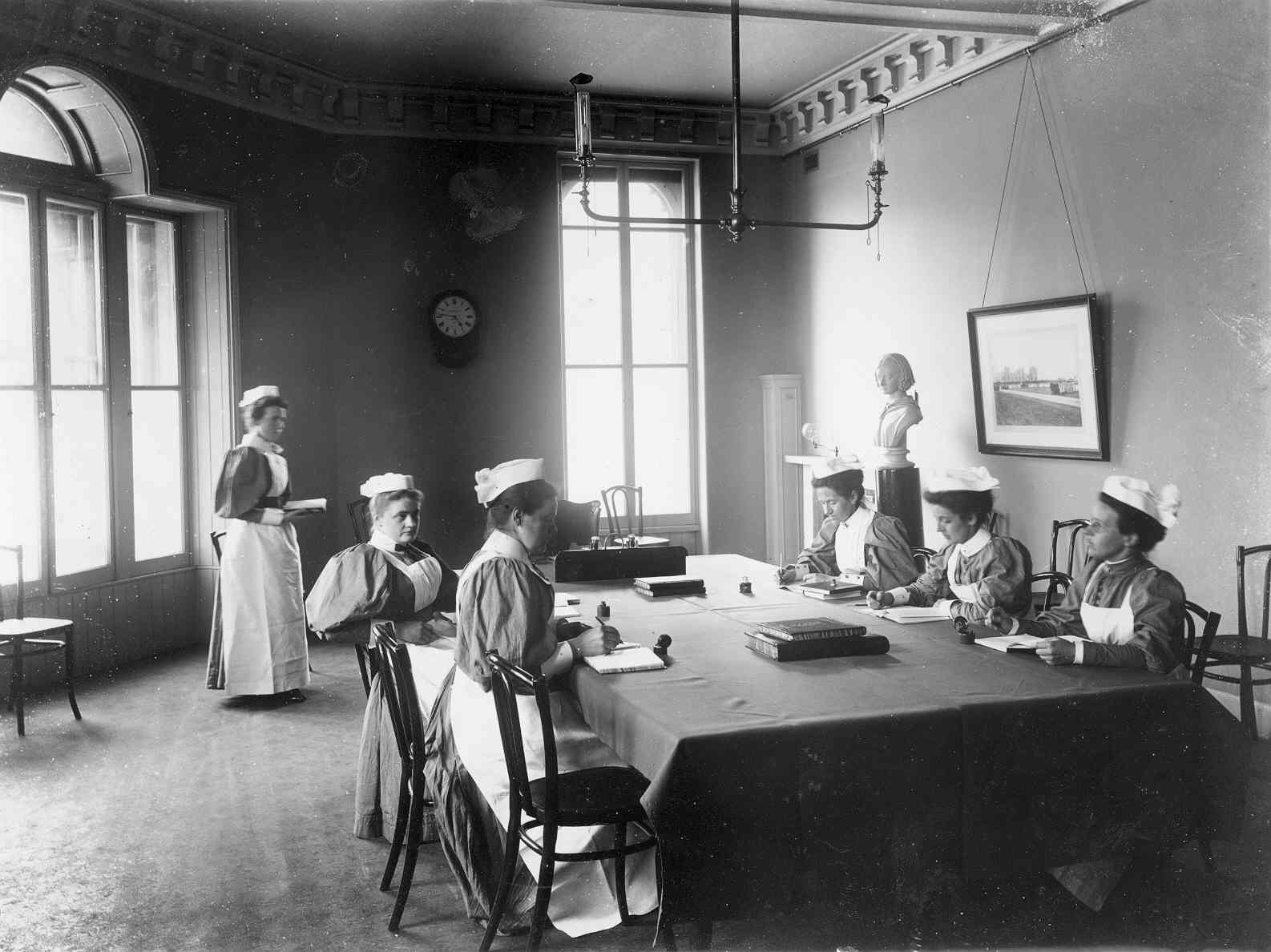 The move to Castle Terrace A black and white image of six people in a large room. Five people sit at a large table in the centre of the space, one person stands. They all wear traditional nursing uniforms, long dress, light-coloured apron and white bonnet-style hat.