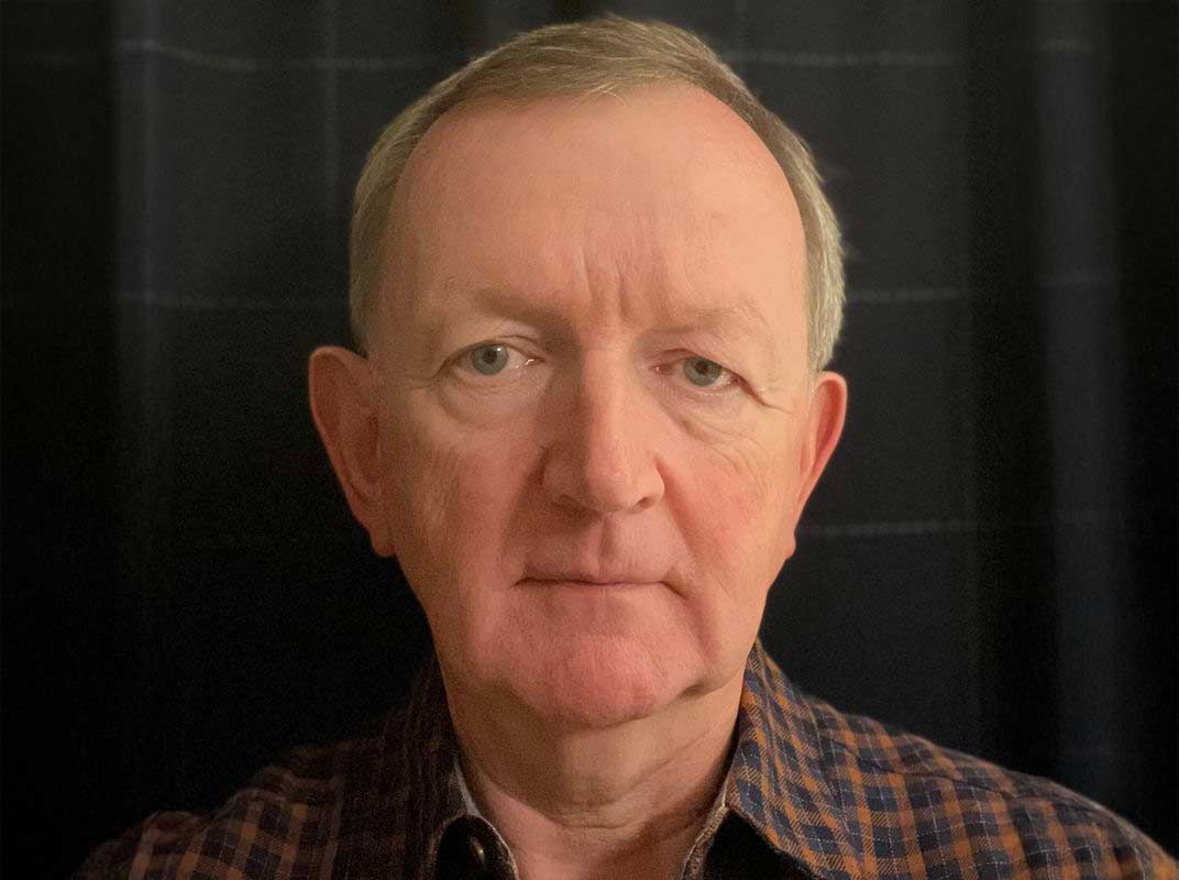 A serious person with short grey hair. They wear a black and orange plaid shirt and stand against a black background.
