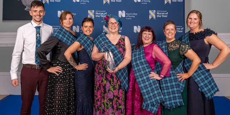 Group of seven people standing in front of a backdrop for the RCN Scotland Nurse of the Year Awards. They are dressed in formal attire, many wearing blue tartan sashes, and one person at the canter holds a glass award. All are smiling and all but one are posing with hand on hip.