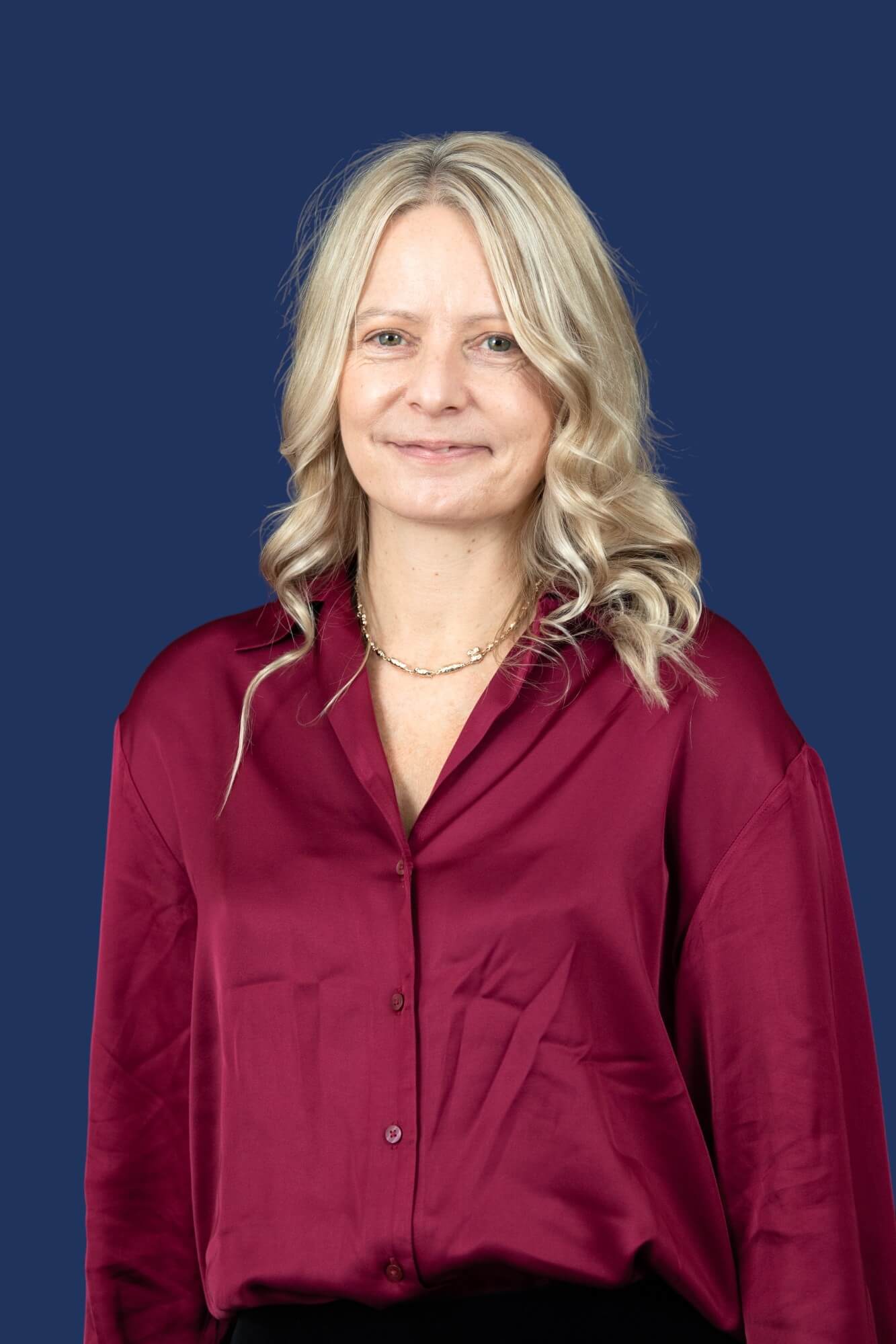 A person with long, wavy light hair is standing against a solid dark blue background. They are wearing a dark red button-up shirt and a gold necklace, and they are looking at the camera with a slight smile.