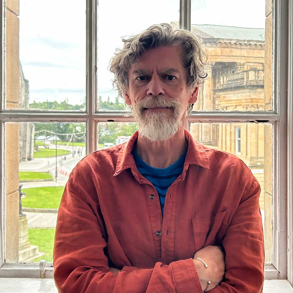 A person with wavy grey hair and a full beard stands indoors with arms crossed, wearing a rust-coloured button-up shirt over a blue striped top. They are positioned in front of a window, with historic stone buildings and a green lawn visible outside.