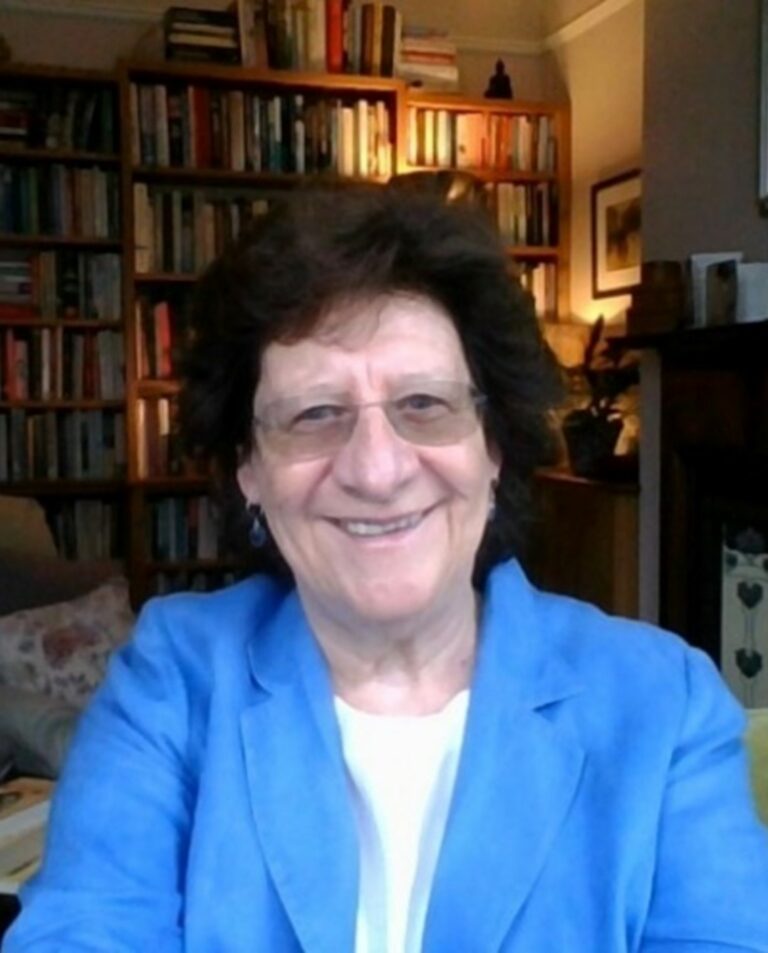 A person with short, dark-coloured hair and glasses, wearing a blue jacket over a white top, stands in front of a bookcase. They are smiling and looking directly at the camera.