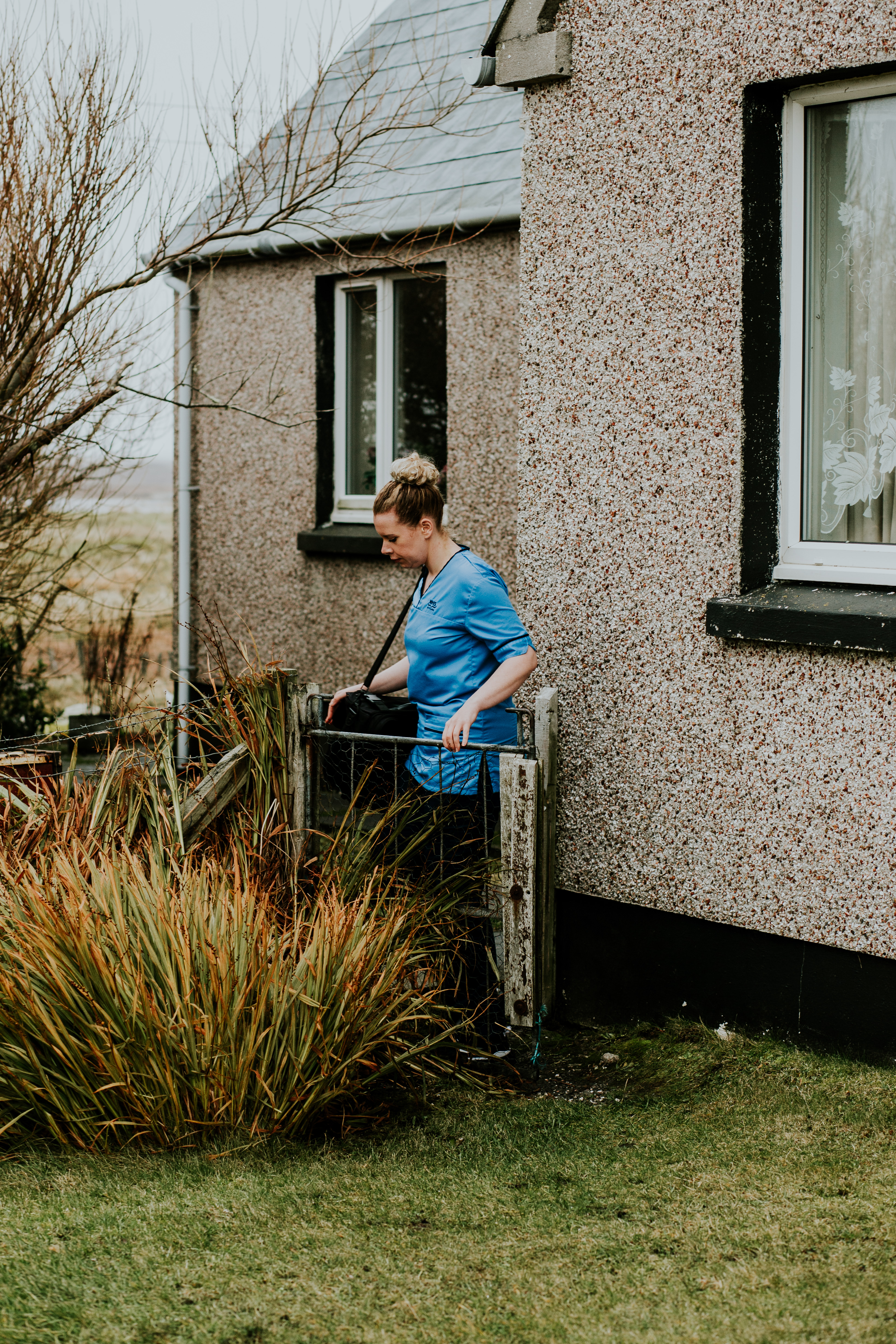 A person standing at an open garden gate with bushes and grass around them and a house wall with windows and roof visible behind them. They are wearing a blue nurse’s uniform – blue top and dark trousers, with a blonde bun and carrying a black bag with a long handle.