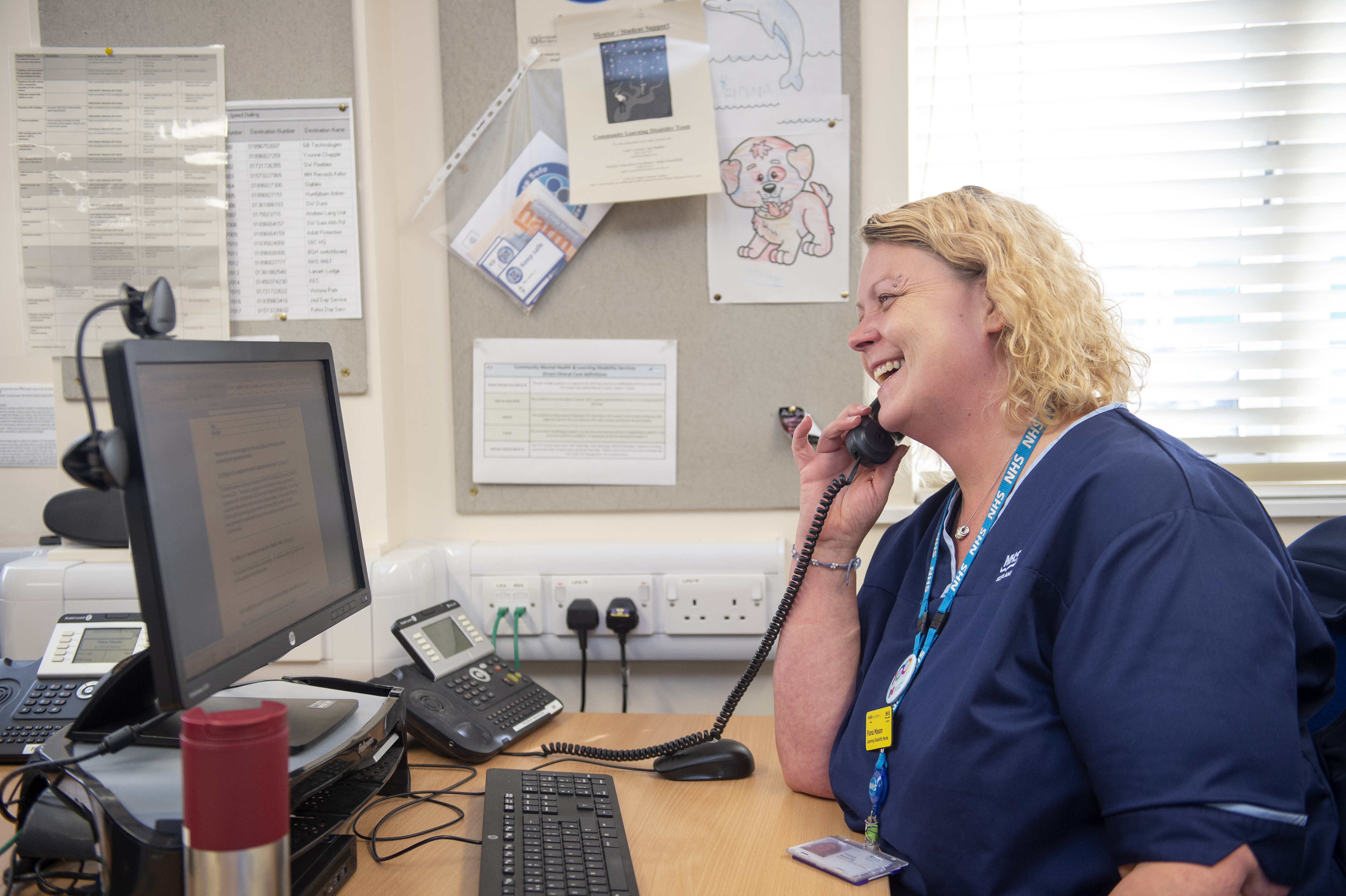 A smiling person with curly blonde hair to their shoulders. They wear a dark blue nurse’s uniform and a blue lanyard with a yellow badge on it and are talking on the phone. They sit at a brown desk with a keyboard, telephone and monitor on it. On the wall next to them are multiple plug sockets, two plugs, two green cables and papers pinned to a pale grey noticeboard.