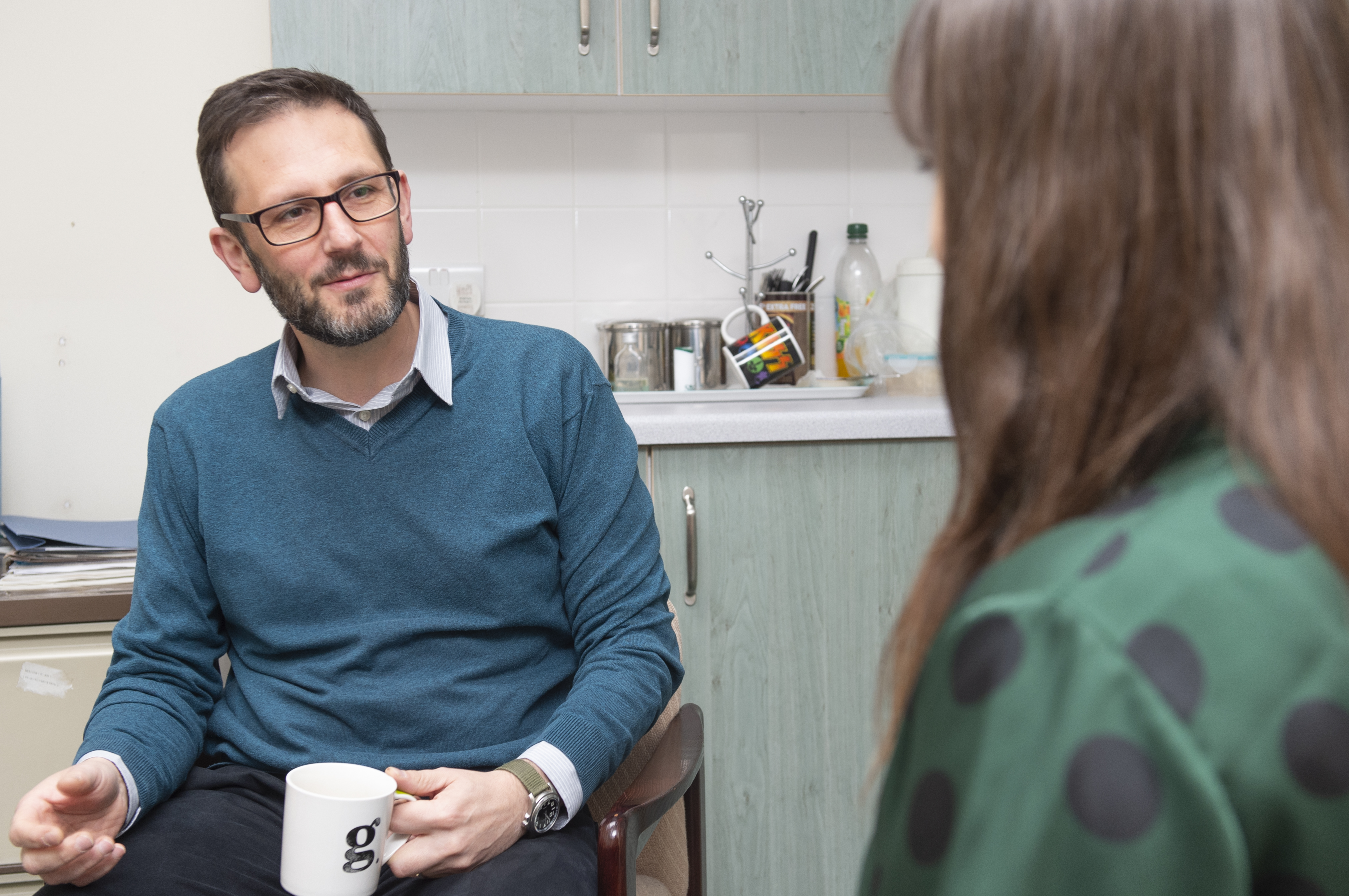 A person with short hair, a beard, and glasses sits in a kitchen or break room holding a white mug with the letter 'g' on it. They are wearing a teal sweater over a collared shirt and are engaged in conversation with another person whose back is to the camera. The second person has long hair and is wearing a green top with large black polka dots. In the background, there is a countertop with mugs, utensils, and a plastic bottle.