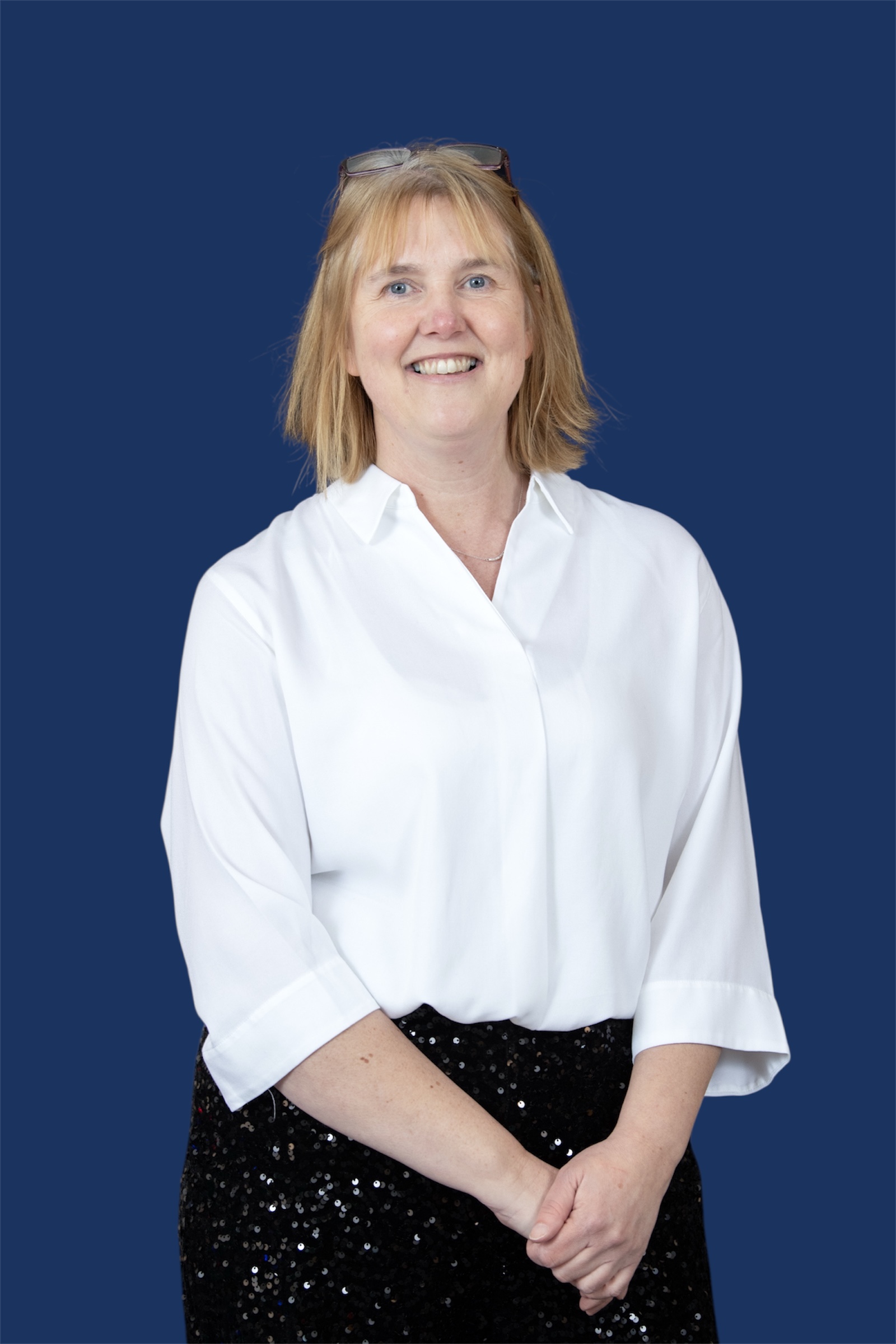 A person with straight, shoulder-length blonde hair stands against a blue background. They are smiling and wear a white, long-sleeved blouse tucked into black, sequined trousers. They have glasses resting on top of their head and their hands are clasped in front of them.