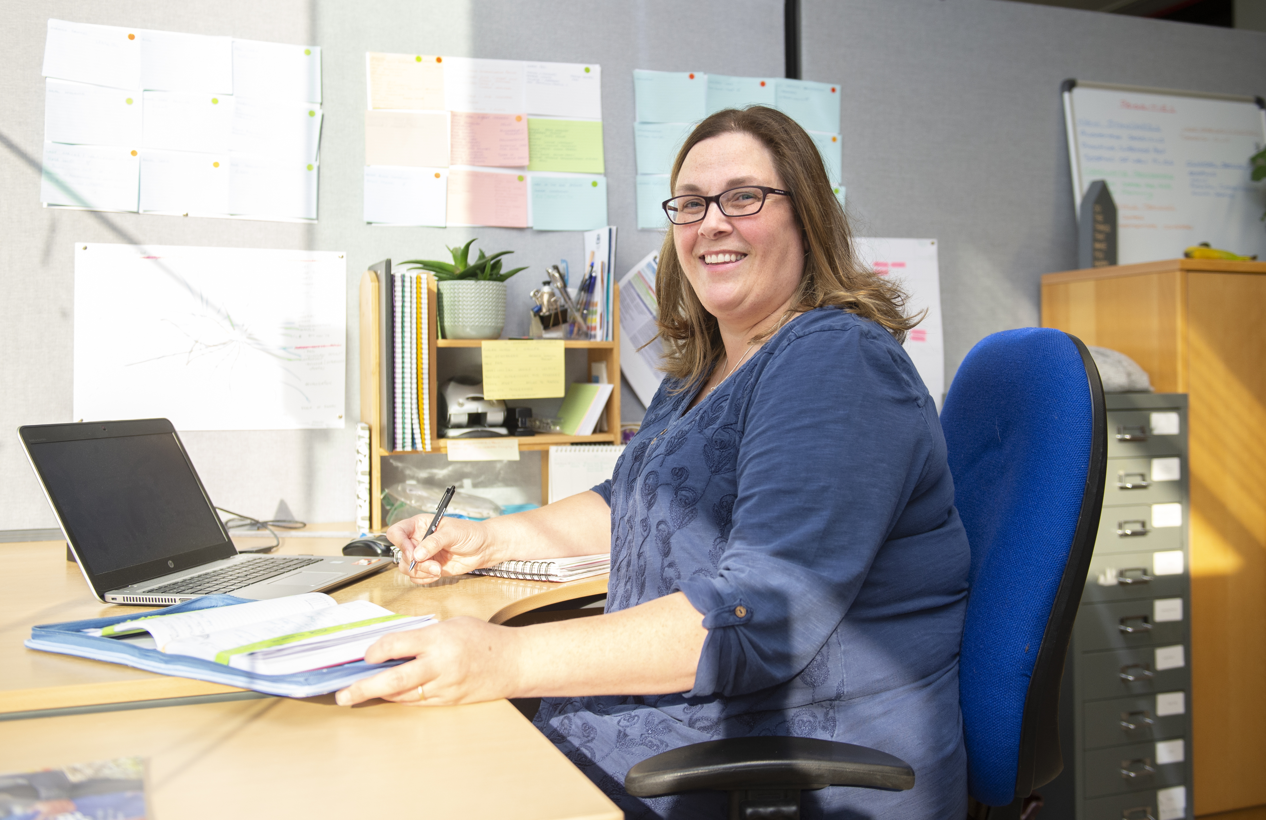A person with medium-length hair and glasses sits at a desk in a brightly lit office. They are smiling at the camera while holding a pen and writing in a notebook. An open folder with papers and a laptop are on the desk. Behind them is a small bookshelf with office supplies, a potted plant, and colourful notes pinned to a bulletin board. Sunlight streams into the room, casting shadows across the workspace.