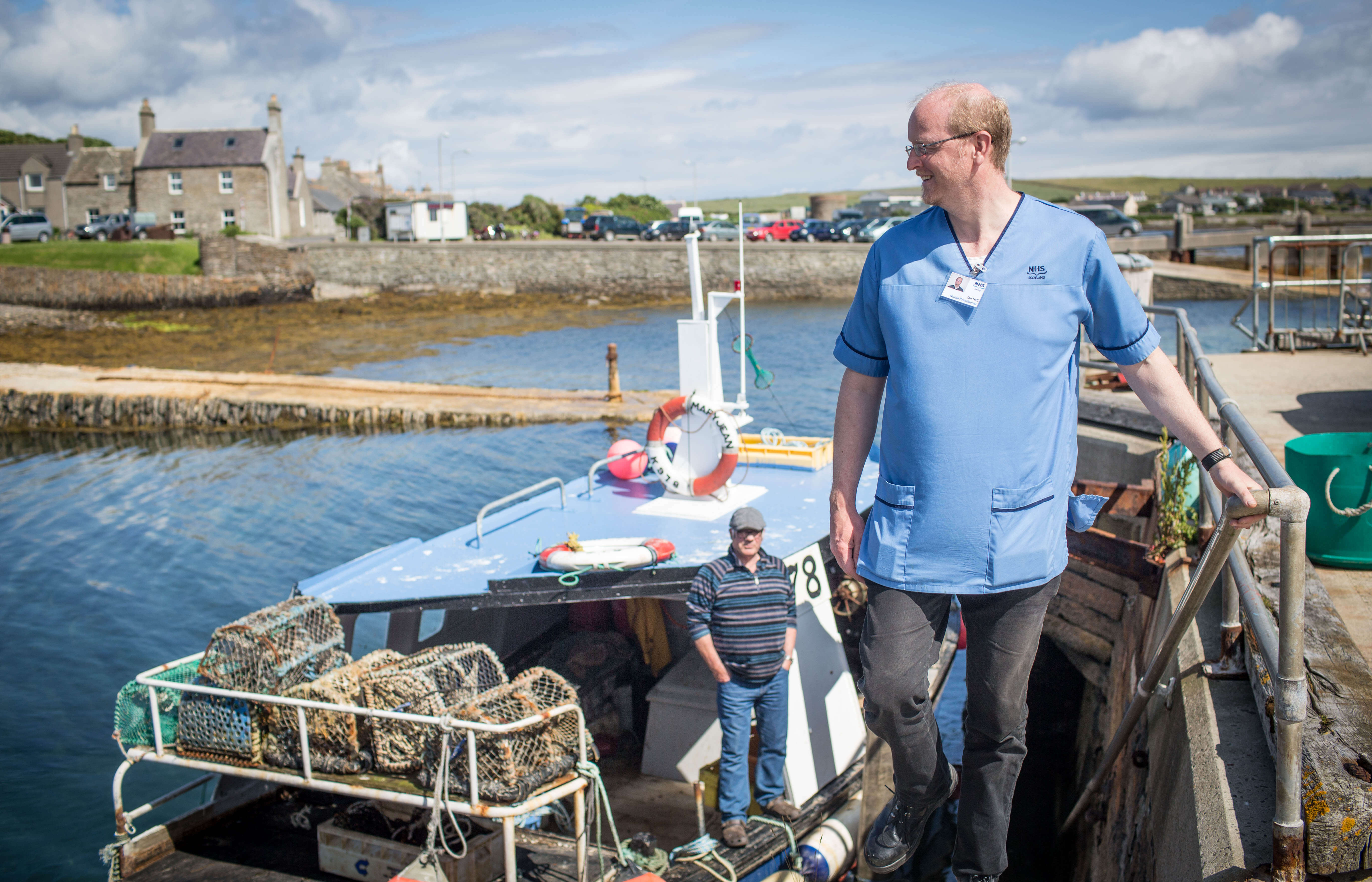 A nurse wearing a blue NHS uniform, glasses and grey trousers smiles while standing on a staircase at a harbour, holding a metal handrail. Behind him, a fisherman stands on a small boat loaded with lobster creels. In the background are stone houses, parked cars, and a partly cloudy sky.