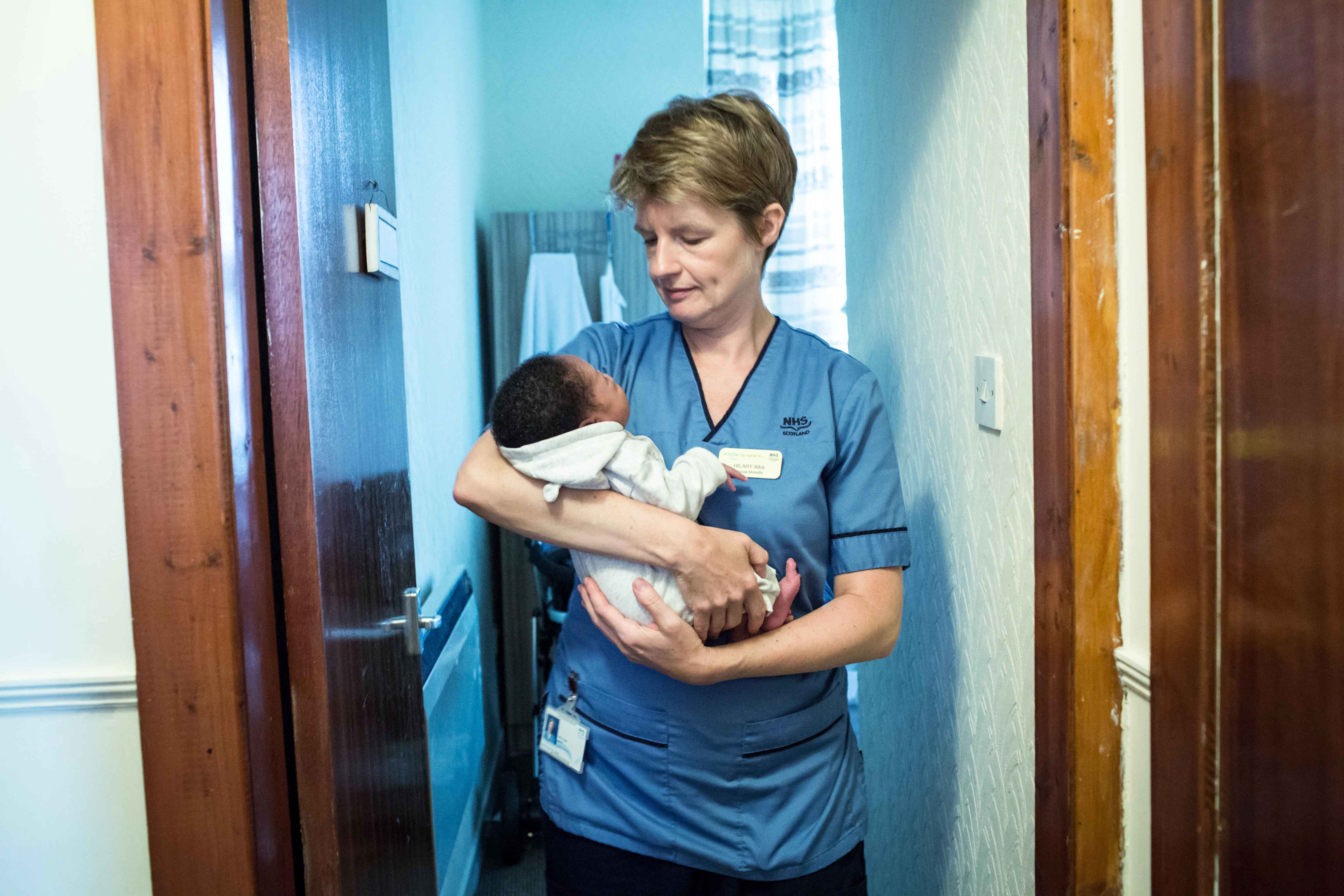 A nurse with short dark blonde hair and wearing a blue NHS uniform stands cradling a black-haired baby dressed in a light grey outfit. The nurse is looking down at the baby. There dark wooden doors to left and right, with a room in the background.