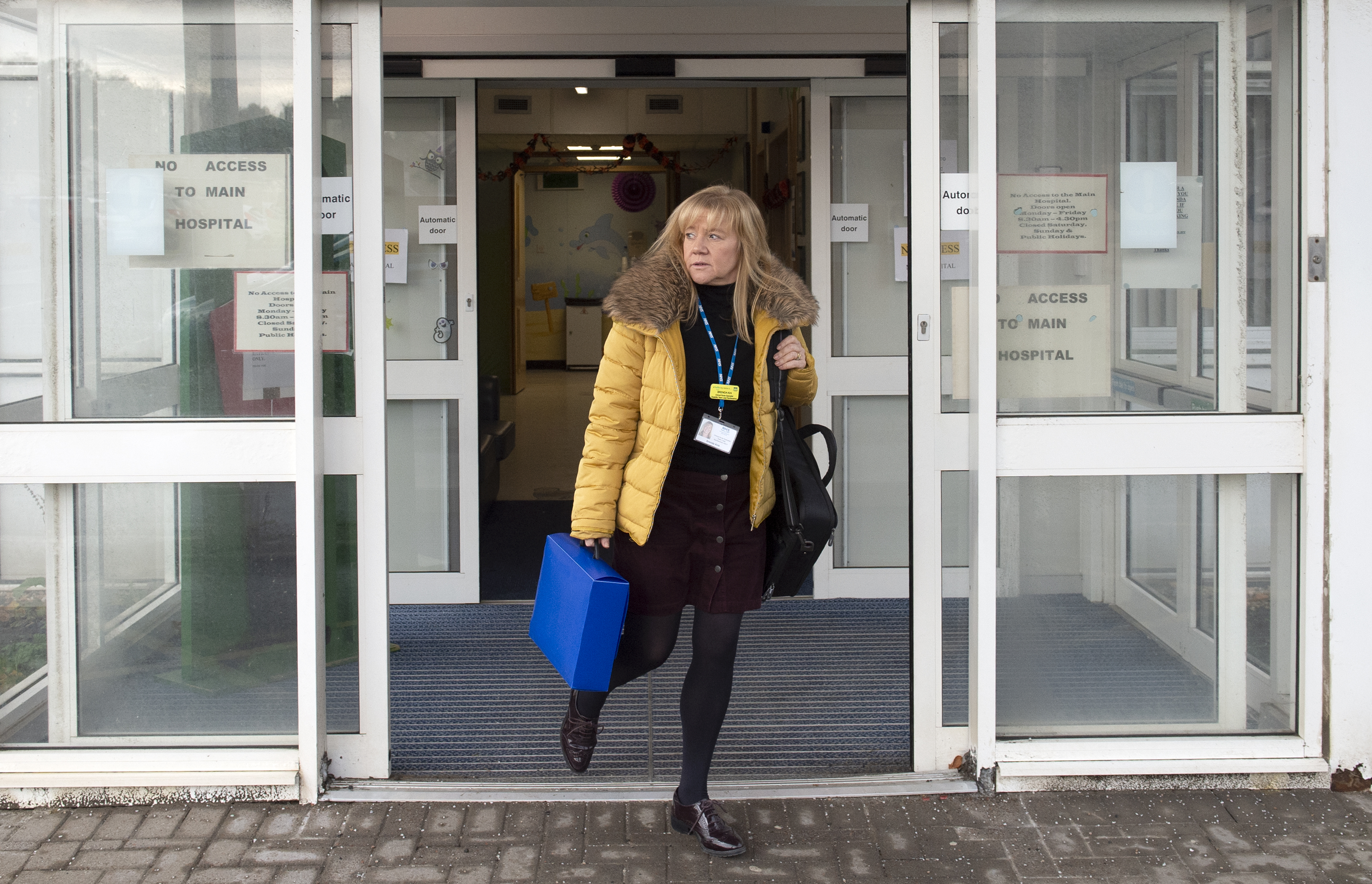 A person with long light-coloured hair, dressed in black apart from a yellow coat walking out of the second of two sliding glass doors. Both sets of doors are open. The person is carrying a black bag over one shoulder and a blue case in their other hand.