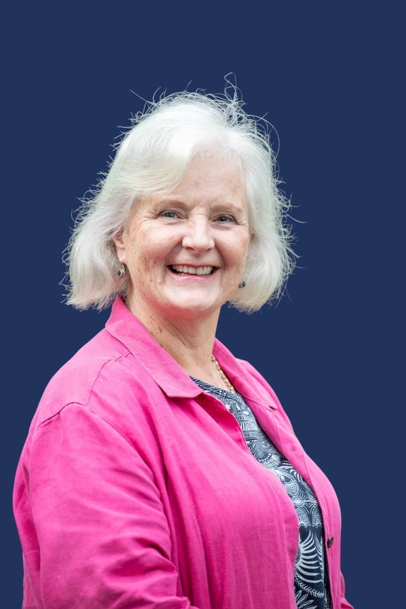 A person with short, wavy white hair is smiling in front of a solid dark blue background. They are wearing a bright pink jacket over a patterned navy and white top.