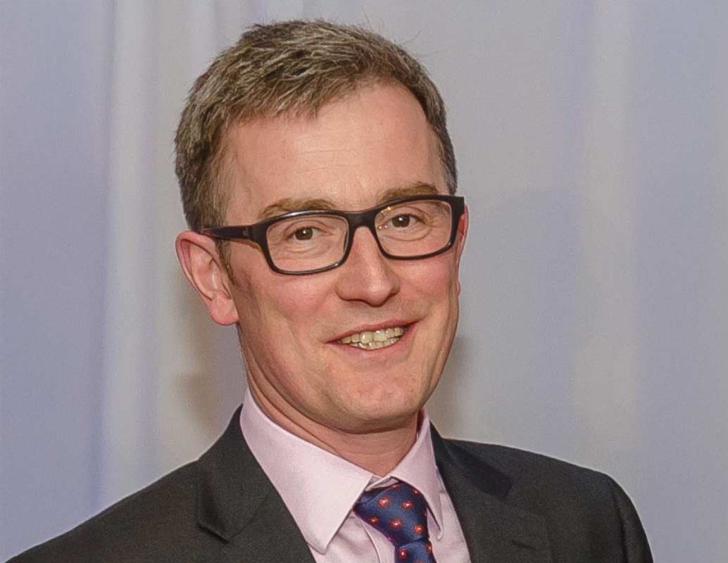 A smiling person in a dark grey suit jacket, pale pink shirt and a blue and red pattern silk tie. They have short grey hair and wear black glasses.