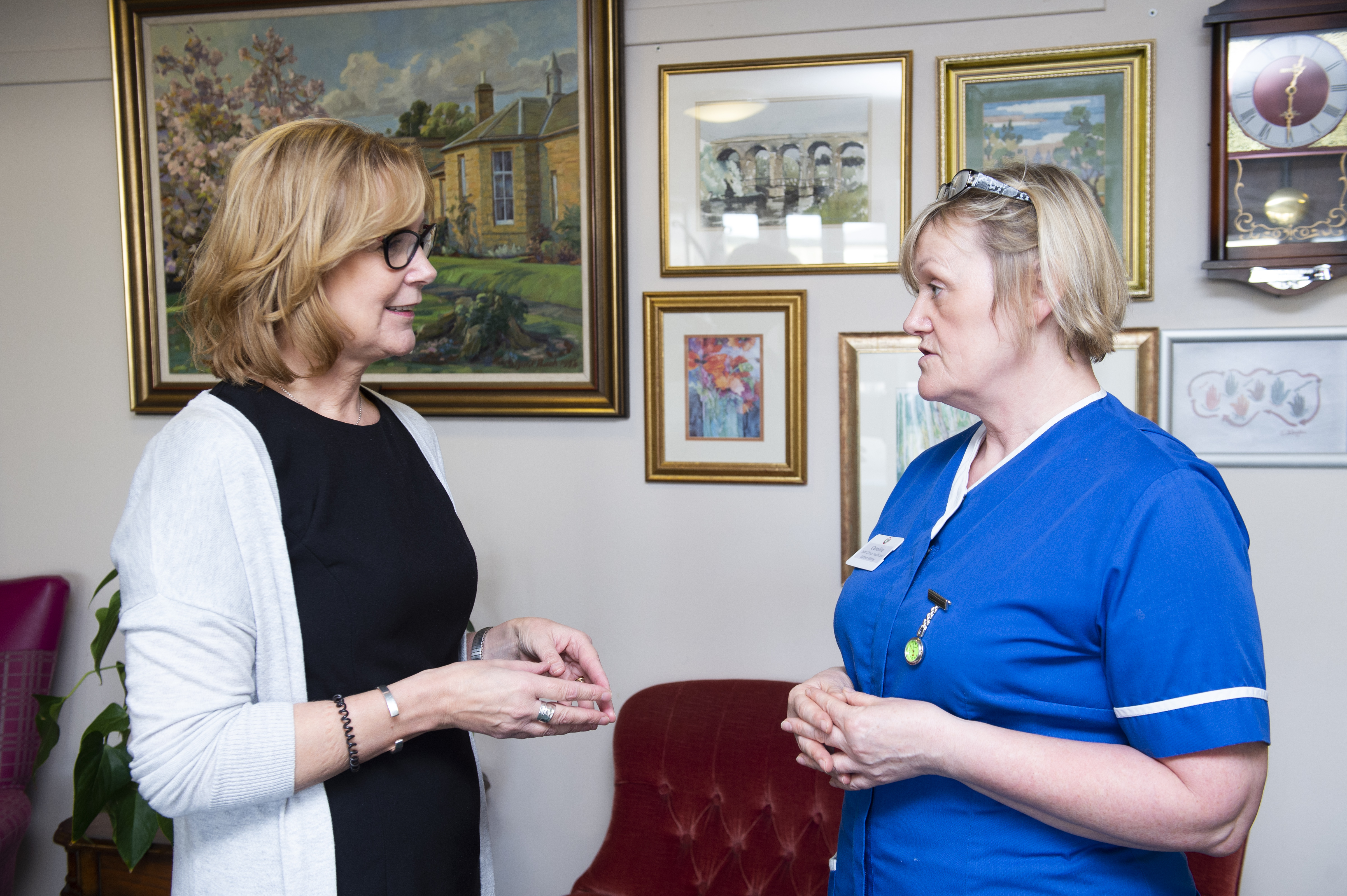Two people are standing and conversing in a room decorated with framed artwork and a large wall clock. One person is wearing a dark outfit with a light cardigan, while the other is dressed in a blue uniform with a name badge and a watch pinned to the chest. They appear to be engaged in a professional or friendly discussion.