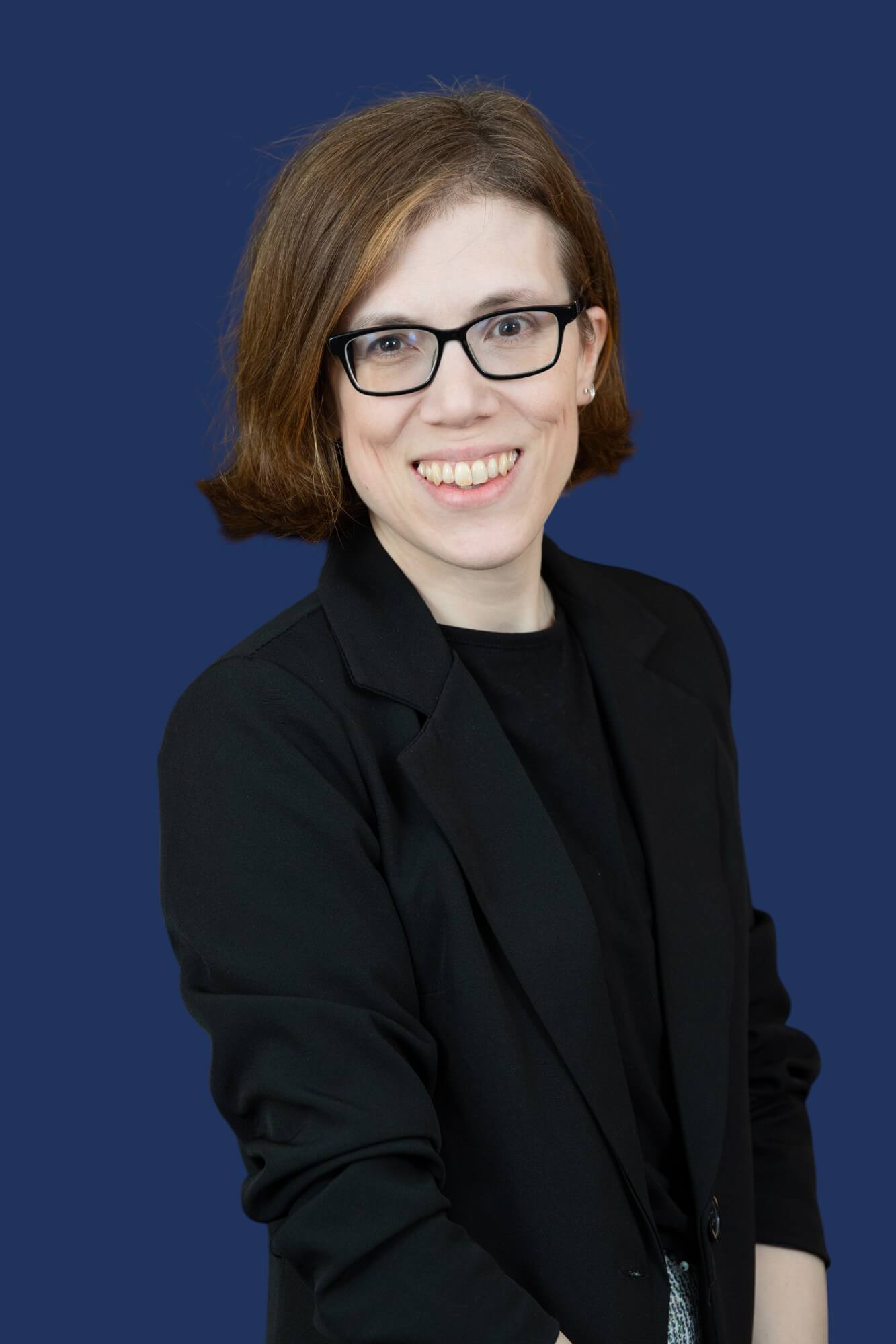 A person with short, light brown hair is standing in front of a solid dark blue background. They are wearing black rectangular glasses, a black blazer over a black top, and are smiling at the camera.