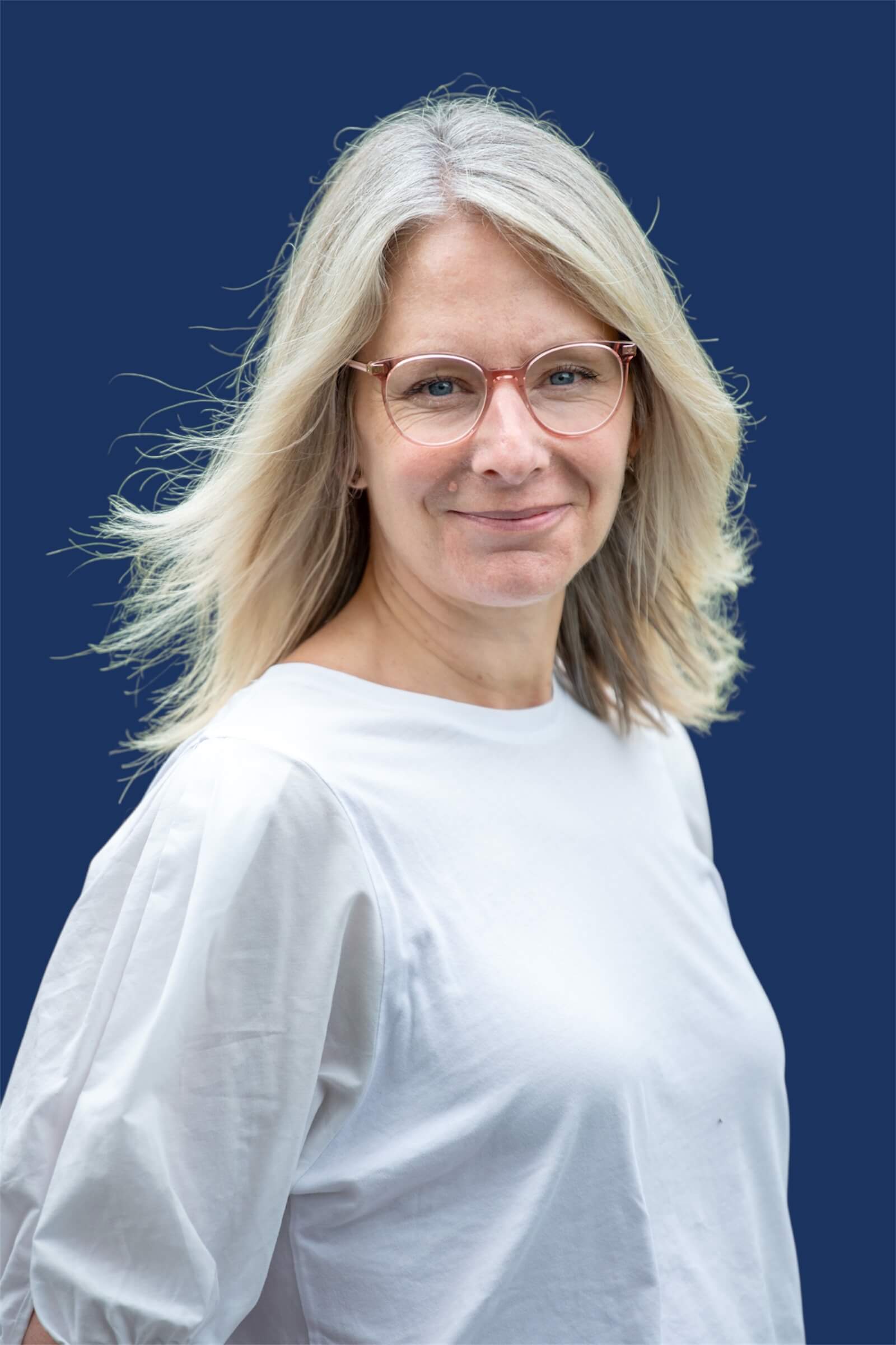 A person with light, shoulder-length hair, wearing glasses and a white shirt, stands in front of a solid dark blue background, looking at the camera with a slight smile.