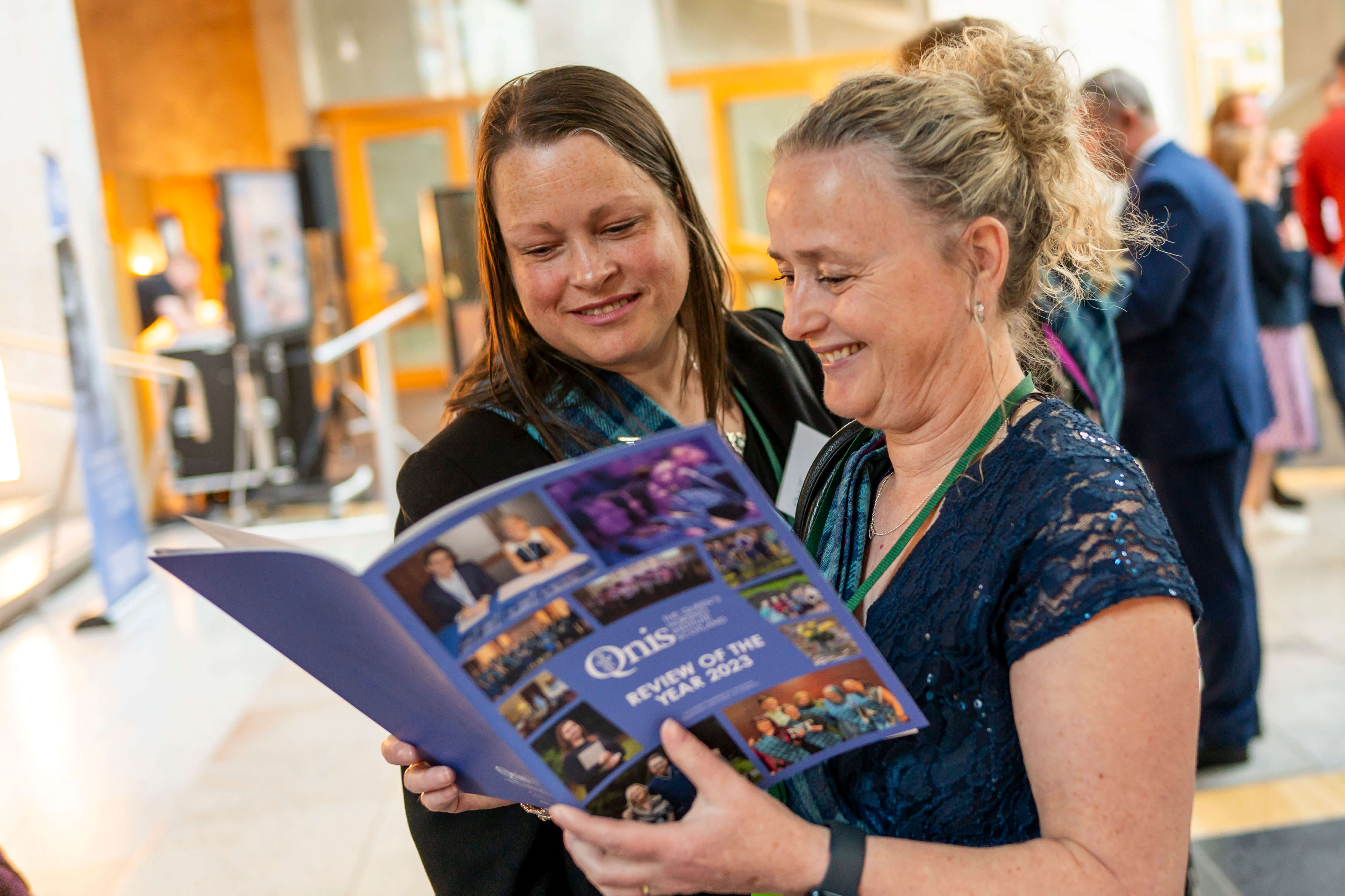Two people standing closely together, smiling as they read a booklet titled 'QNIS Review of the Year 2023'. The background shows other people engaged in conversation in a brightly lit indoor space.’
