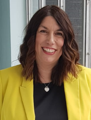 A person with long wavy dark hair, wearing a bright yellow blazer over a black top, smiling while standing indoors near large windows.