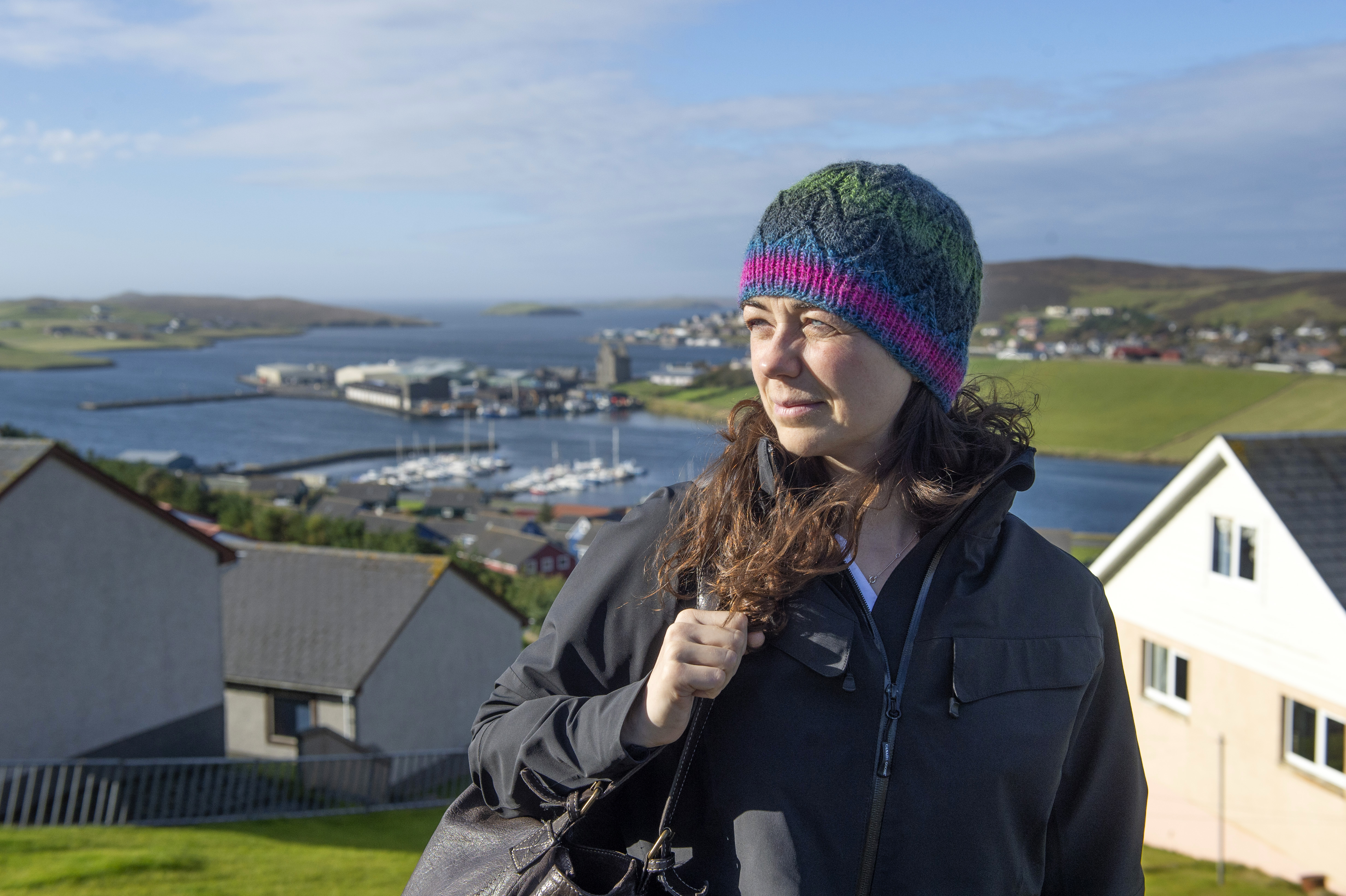 A person with long wavy brown hair wearing a multicoloured knitted hat, deep blue jacket and a backpack on one shoulder. They have sunlight on their face as they look into the distance. In the background blue sky, a large body of water, green hills and houses.
