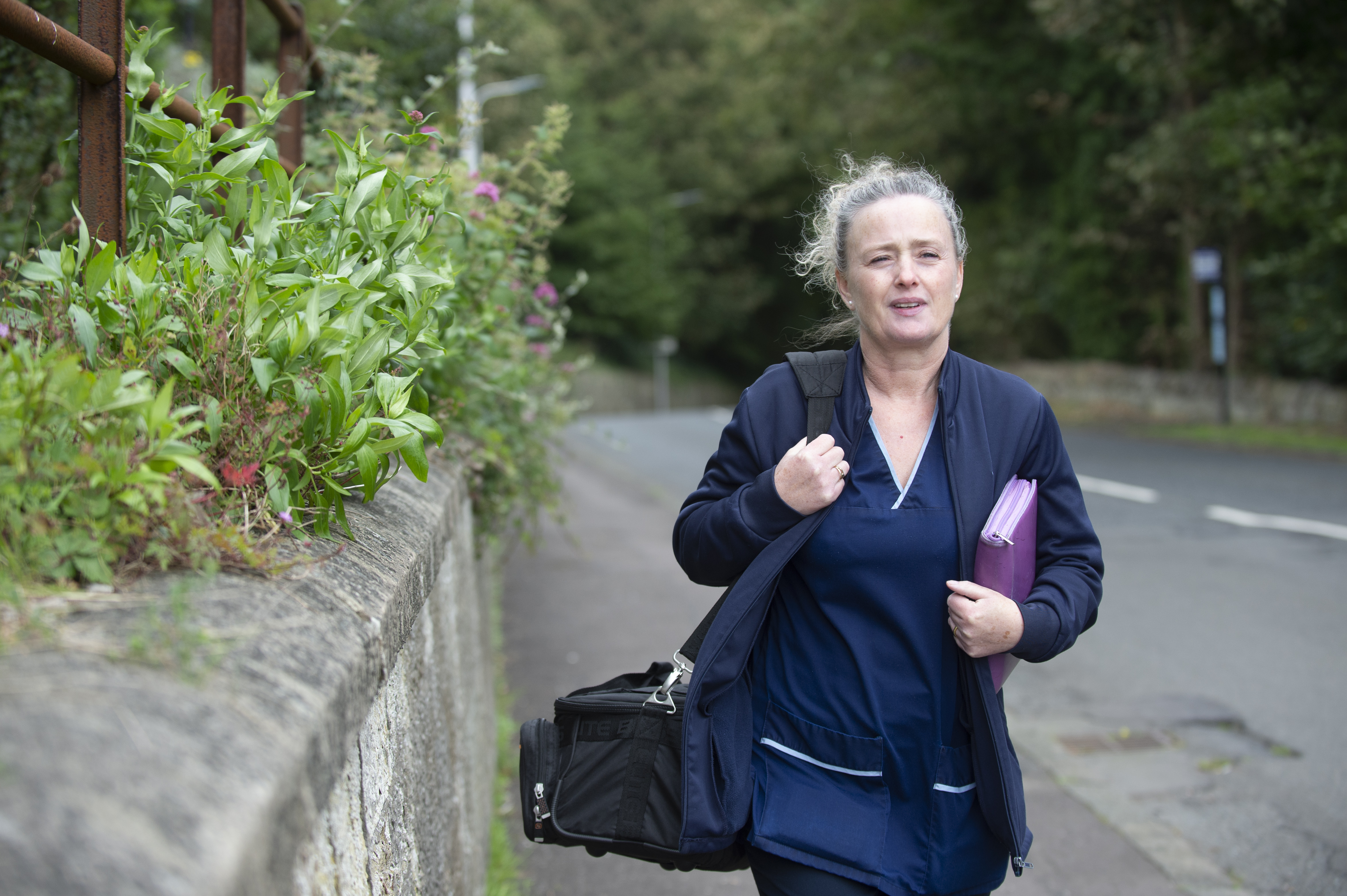 A person wearing a dark blue nurse’s uniform with a deep blue jacket over it. They have light-coloured hair tied in a ponytail and carry a black bag on one shoulder and a purple folder under their other arm. They walk on a grey pavement with a wall on one side covered with greenery, and a road on the other side.