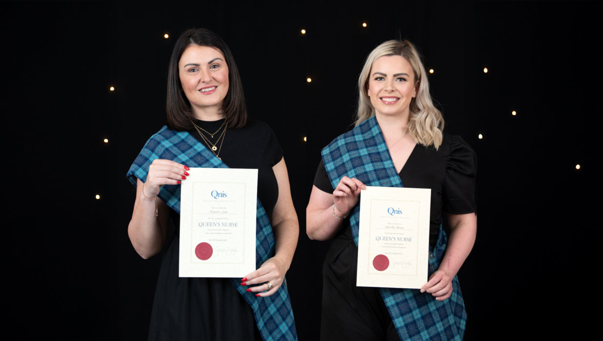 Two individuals stand side by side in front of a black backdrop with small lights, each wearing a blue tartan sash over a black outfit and holding a certificate titled “Queen’s Nurse” from the Queen’s Nursing Institute Scotland (QNIS). Both are smiling and facing the camera.