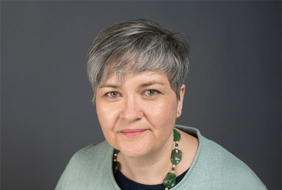A person with short, grey hair styled to the side, wearing a light green sweater over a dark top and a chunky green beaded necklace. They are facing the camera with a slight smile against a plain dark grey background.