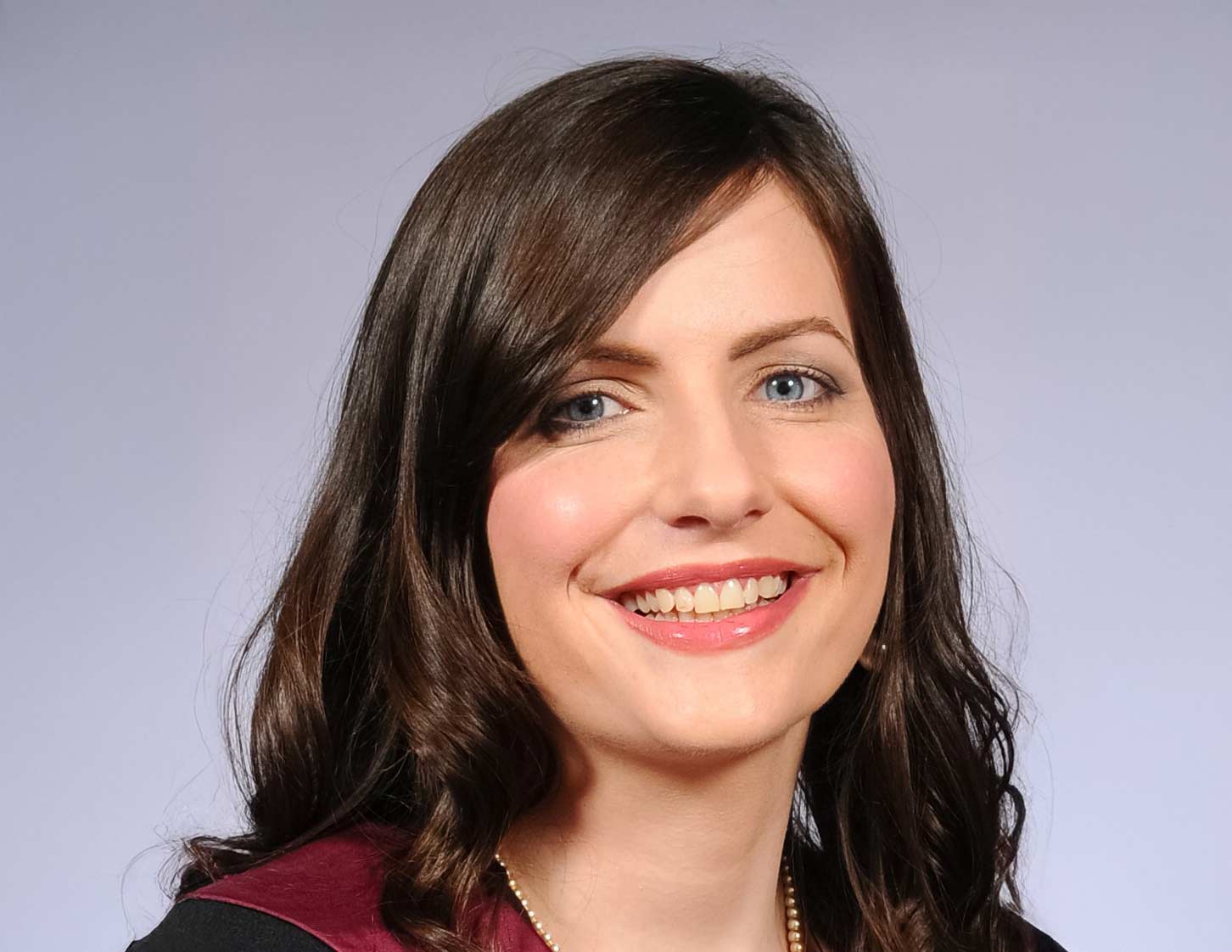 A close-up of a person smiling. They have long dark hair and wear a small pearl necklace. A black and purple top is visible. They are against a grey background.