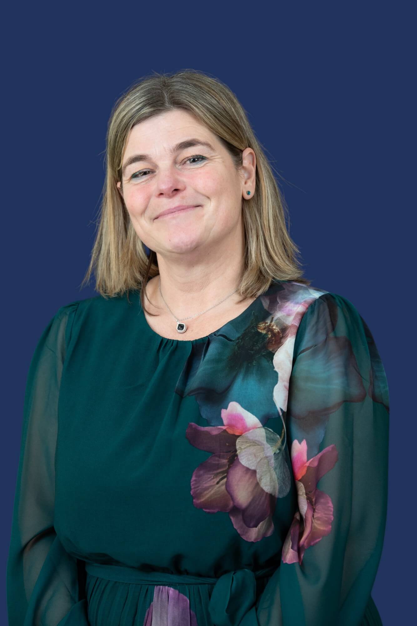 A person with straight, shoulder-length light brown hair is smiling in front of a solid dark blue background. They are wearing a dark green top with semi-sheer sleeves and a large, colourful floral print.