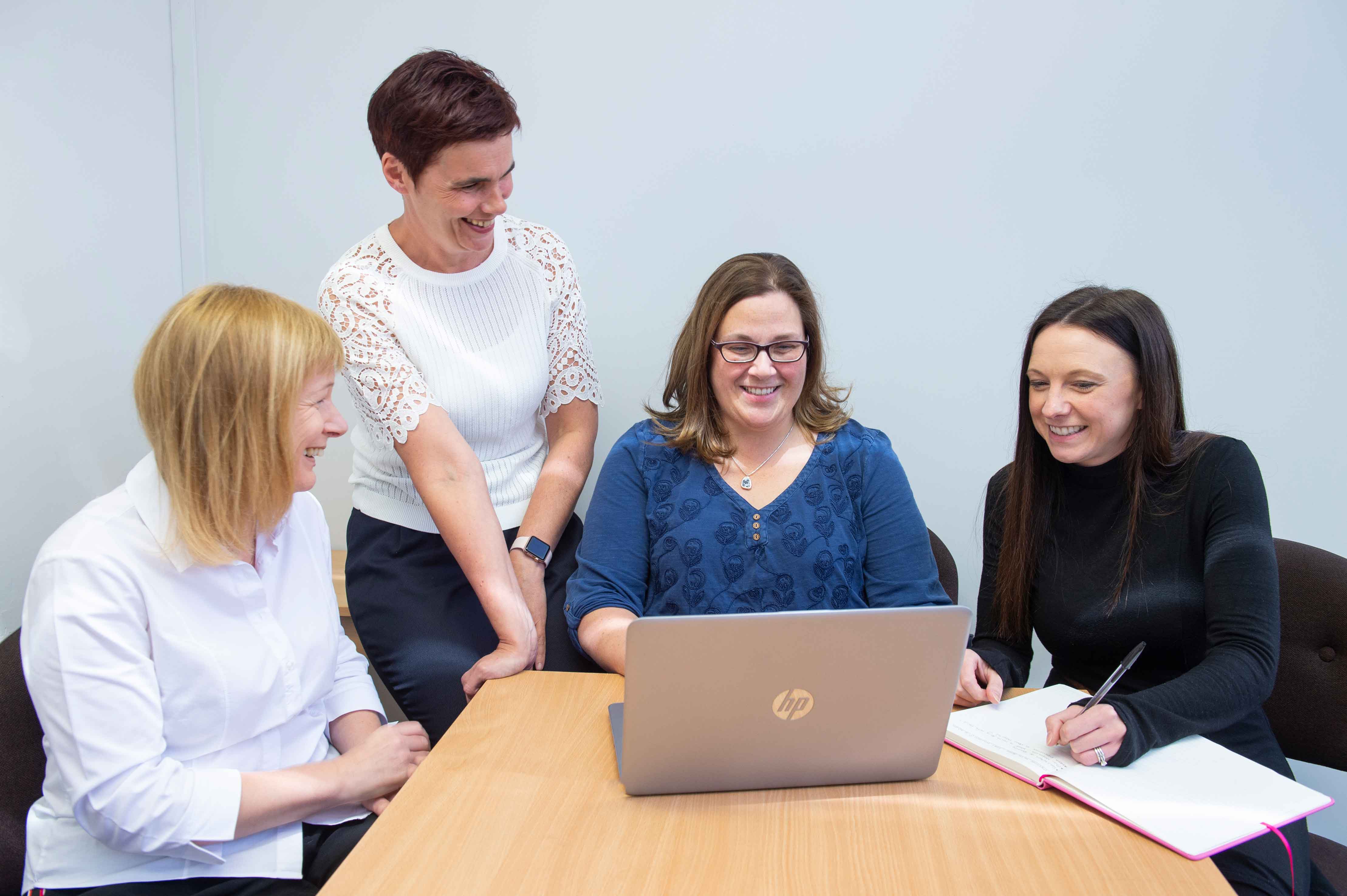 Four people sitting and standing around a light wooden table, smiling and engaged in discussion. One wearing glasses and a blue top is seated and working on an open HP laptop. Another in a black top is writing in a notebook. Two others, one in a white shirt and one in a white top with lace sleeves, are smiling and looking at the laptop screen. The setting is a bright, modern room with a plain light grey wall.