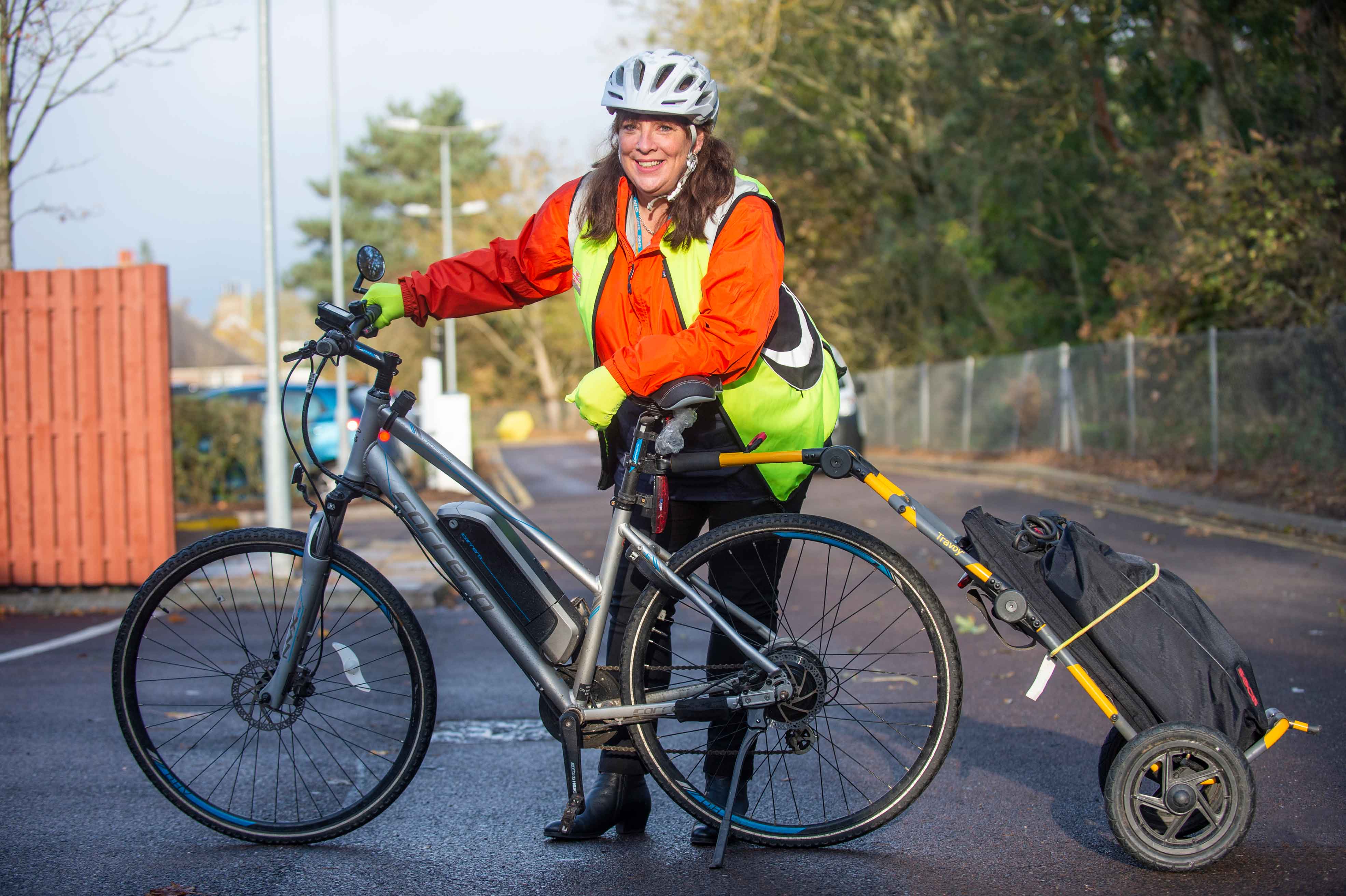 A smiling person with long dark hair wearing a white helmet, fluorescent yellow vest, and orange jacket standing next to a bicycle. The bicycle has a small trailer attached, carrying a black bag. They are outdoors on a paved road lined with trees and a red fence on one side and a silver fence on the other.