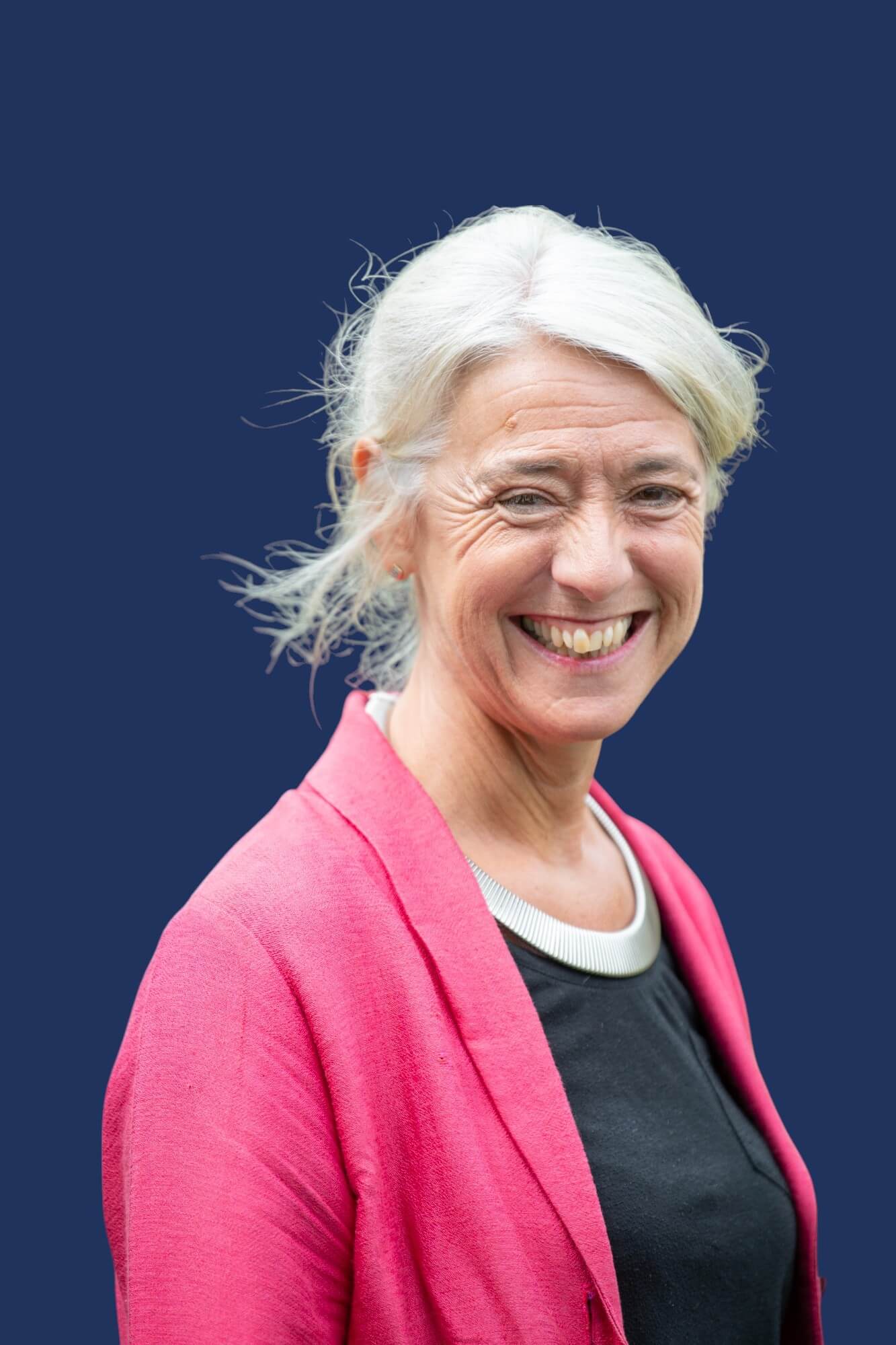 A person with white hair tied back is smiling while looking toward the camera. They are wearing a black top with a white collar detail and a bright pink outer garment, set against a solid dark blue background.