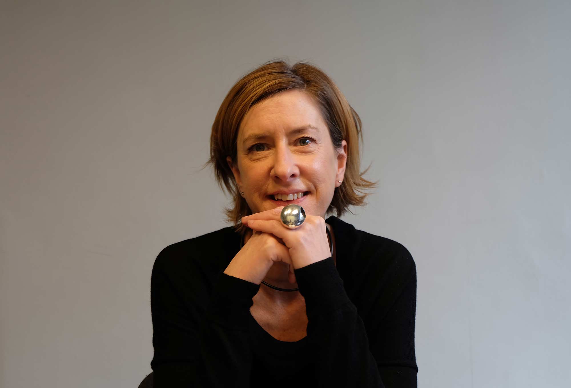A person smiling with hands on chin. They have short brown hair and wear a black round neck long-sleeved top and a silver ring on one hand, and sit against a cream background.