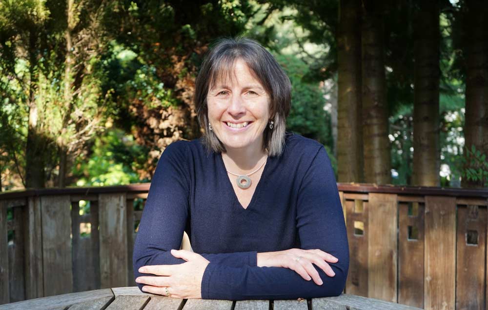 A person with straight, shoulder-length brown hair sits at a round wooden outdoor table, smiling warmly. They are wearing a dark long-sleeved top and a circular pendant necklace, with trees and wooden fencing in the background.
