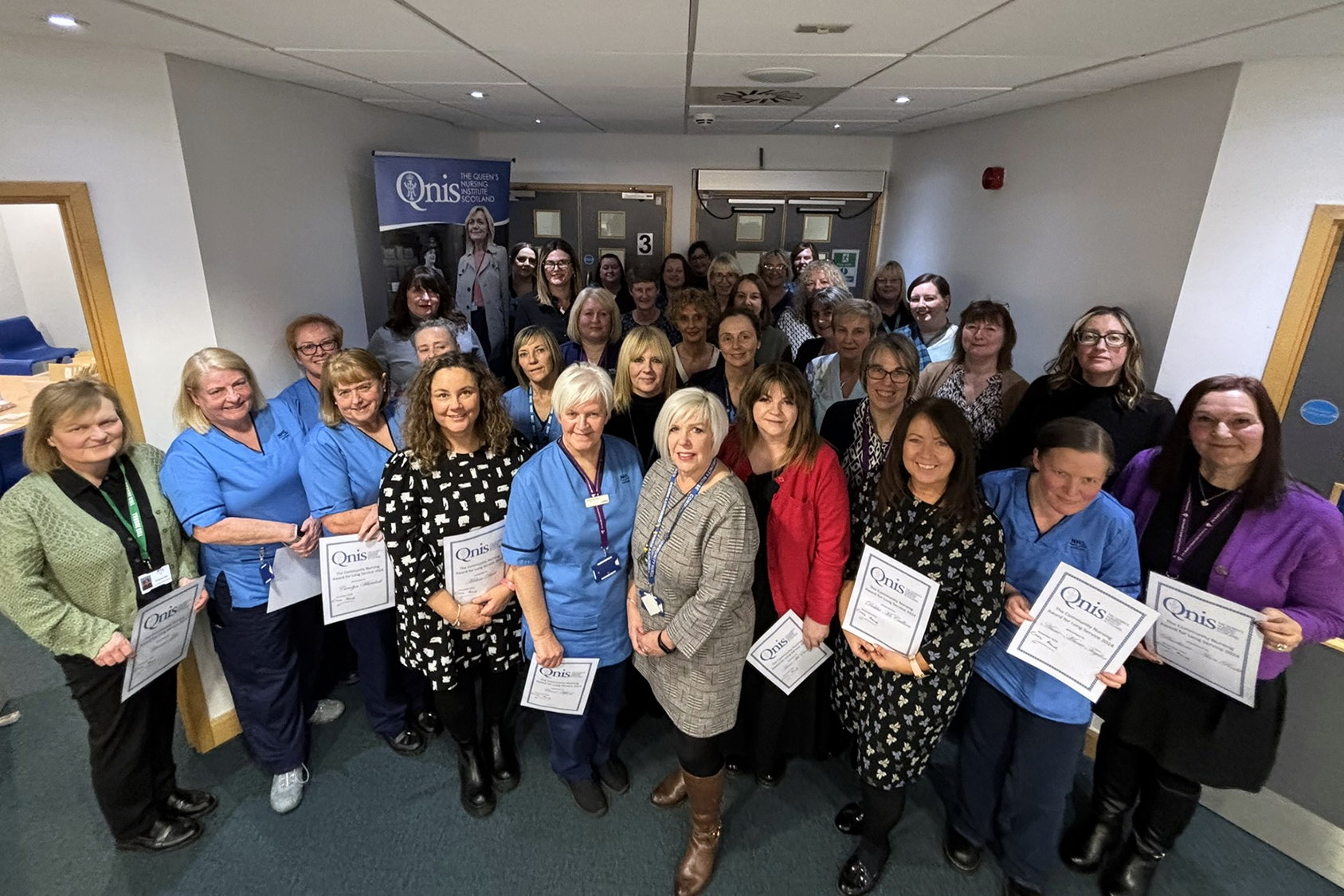 A large group of people pose together in a hallway, smiling toward the camera. Several individuals at the front are holding certificates with the QNIS (Queen’s Nursing Institute Scotland) logo, suggesting an award or recognition event. Some are wearing blue uniforms, while others are dressed in professional or casual clothing. A QNIS banner is visible in the background.
