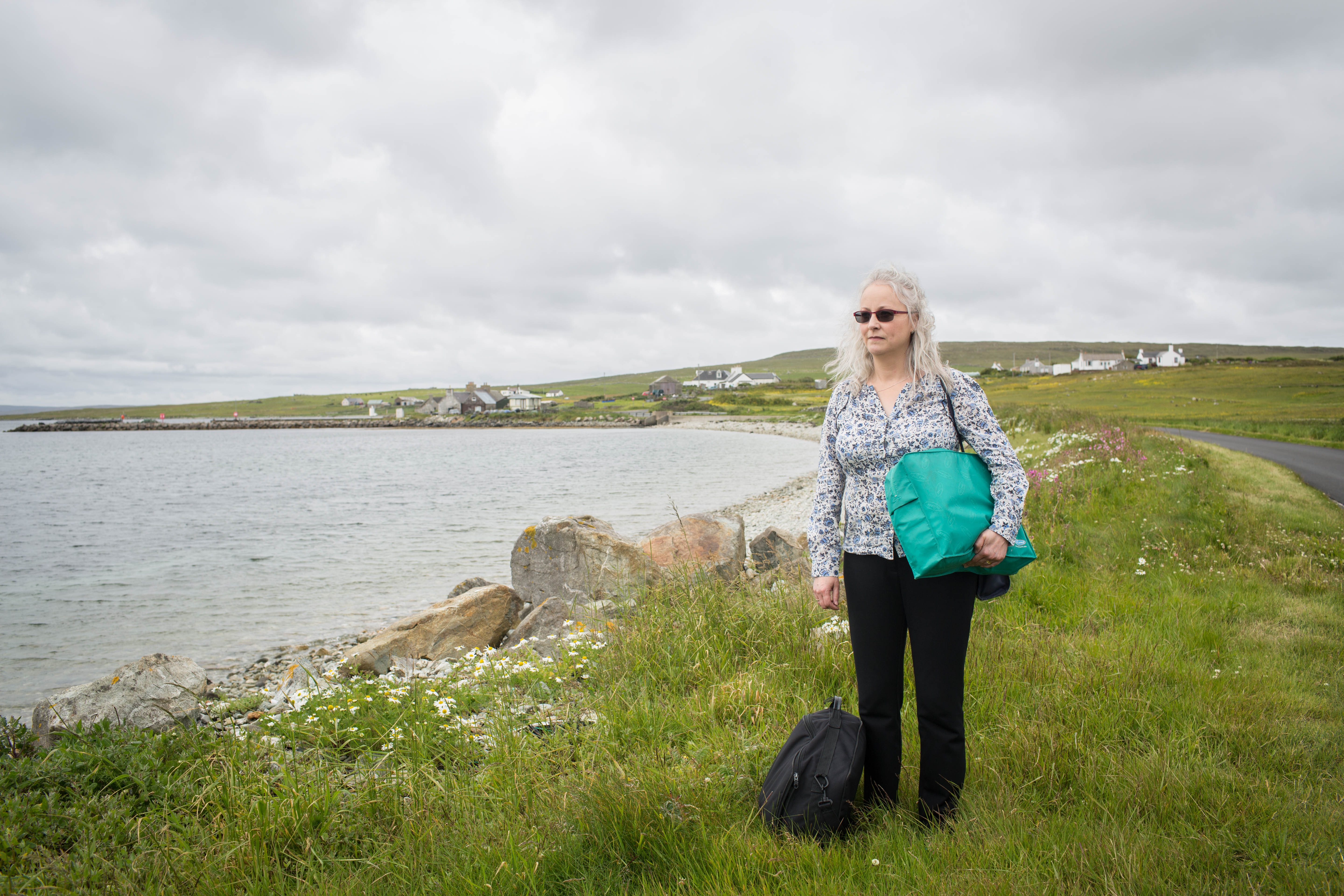 A person with long light hair stands on a grassy shoreline near a rocky beach, holding a bright teal bag and standing beside a black backpack. They are wearing dark trousers and a patterned shirt. The sky is overcast, and small houses are visible in the distance across the bay.