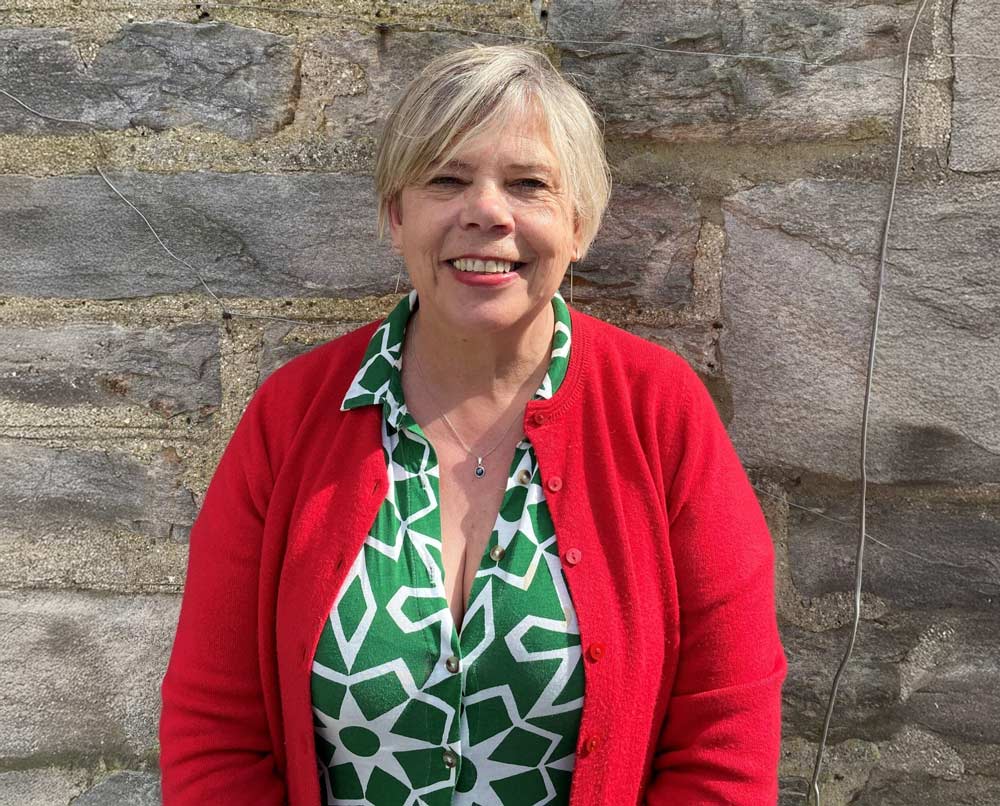 A person smiling in front of a stone wall. They have short light-coloured hair and wearing a red cardigan over a green and white patterned blouse.