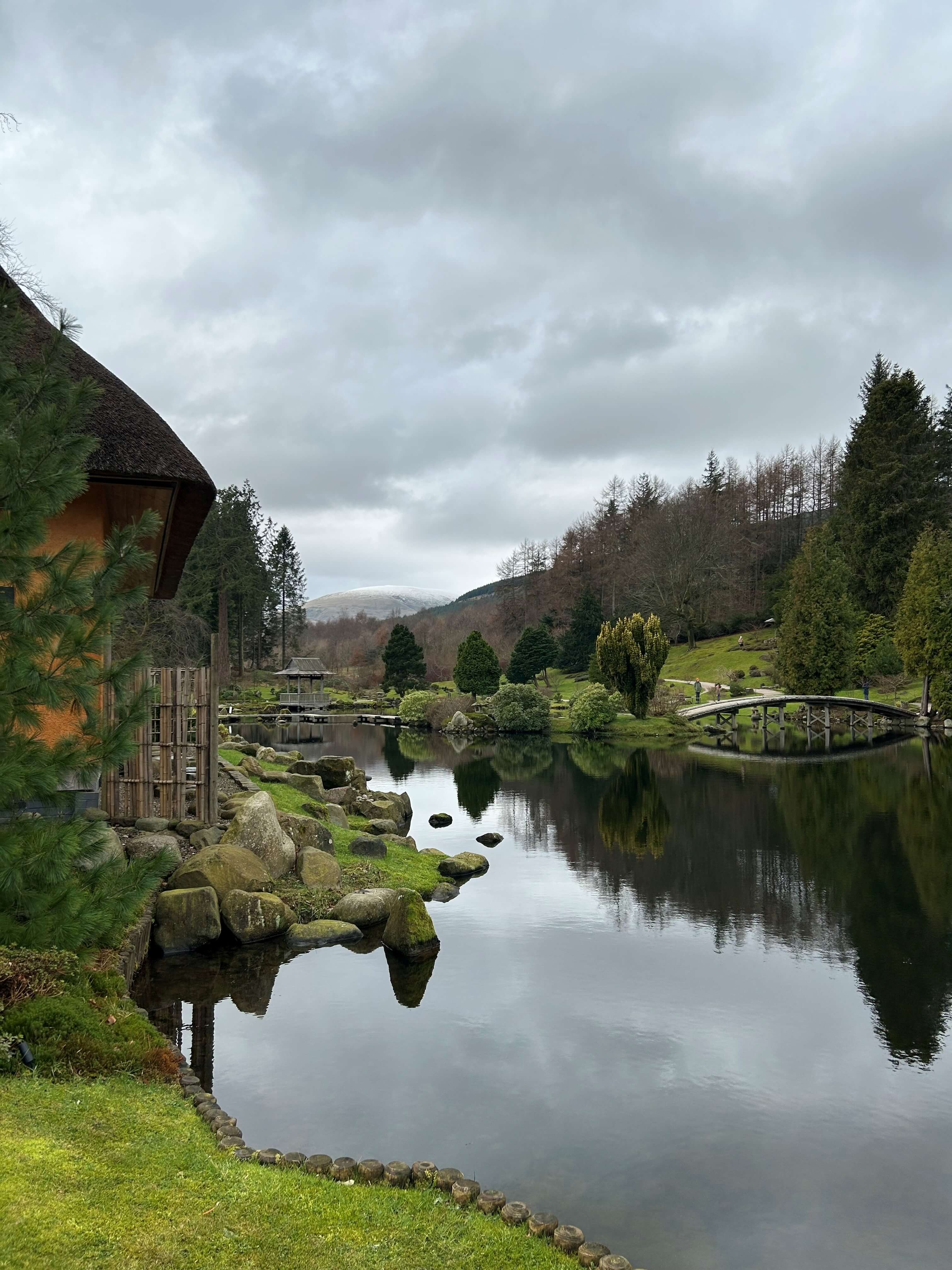 A calm, reflective pond surrounded by green grass, moss-covered rocks, and a variety of trees, with a small wooden bridge crossing part of the water. On the left, a thatched-roof structure is partially visible among tall evergreen trees. In the background, rolling hills and distant mountains are visible under a cloudy, overcast sky.