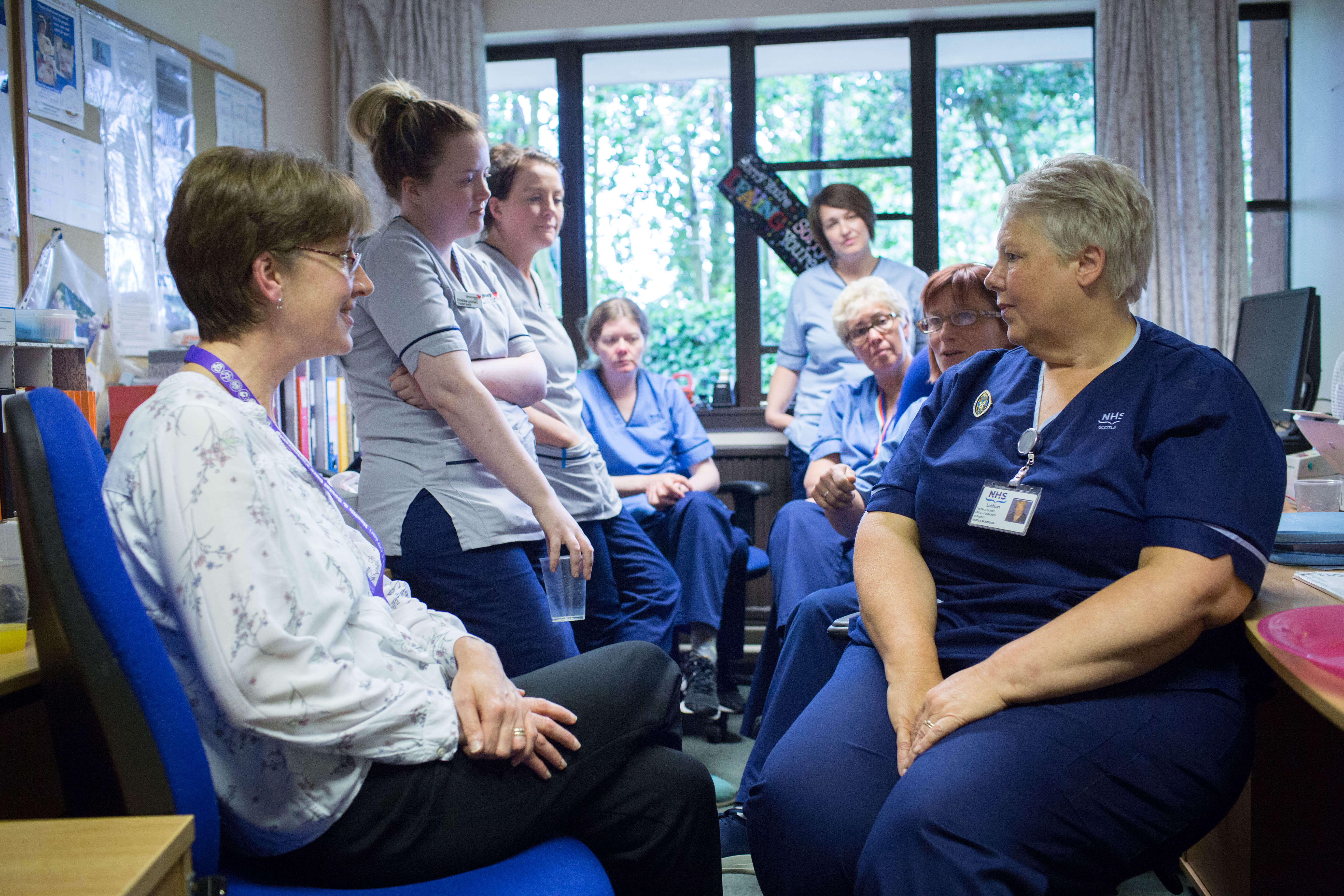 A group of people in an office, some seated and some standing, engaged in conversation. Most are wearing NHS uniforms in shades of blue and grey. In the background is a window showing greenery outside.