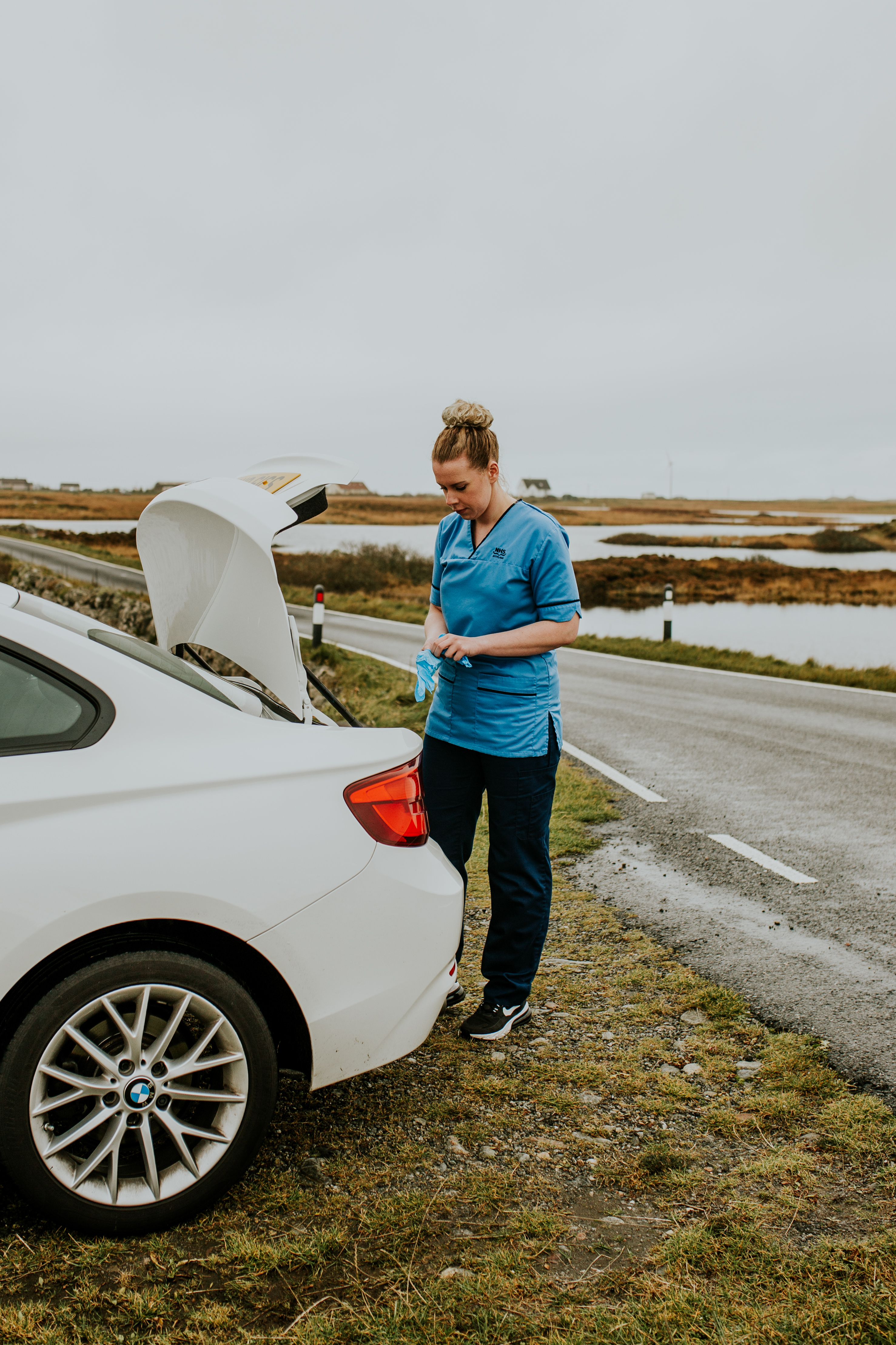 A person wearing a blue nurse’s uniform – blue top and dark trousers, with black trainers and a blonde bun standing next to a white car with the boot open. They are putting on blue latex gloves. There is short grass, water and a road behind them.