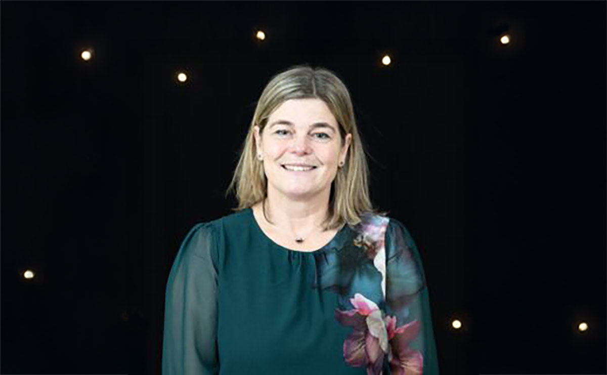 A person with straight, shoulder-length light brown hair is smiling while standing in front of a black background with small lights. They are wearing a dark green top with sheer sleeves and a floral design on one shoulder.