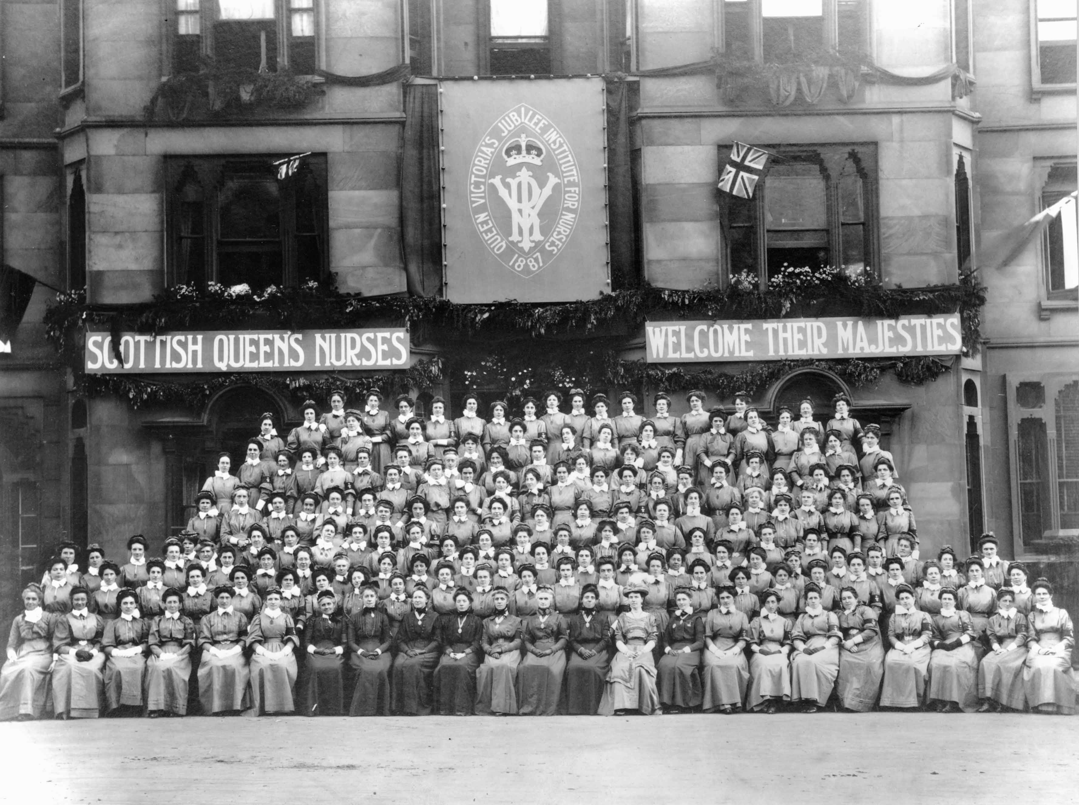 A historic black-and-white photograph showing a large group of people in uniform posed in rows outside a stone building. Banners above them read 'Scottish Queens Nurses' and 'Welcome Their Majesties'. A large emblem in the centre displays 'Queen Victoria’s Jubilee Institute for Nurses 1887'. The individuals are dressed in period nursing uniforms, and some Union Jack flags are visible.