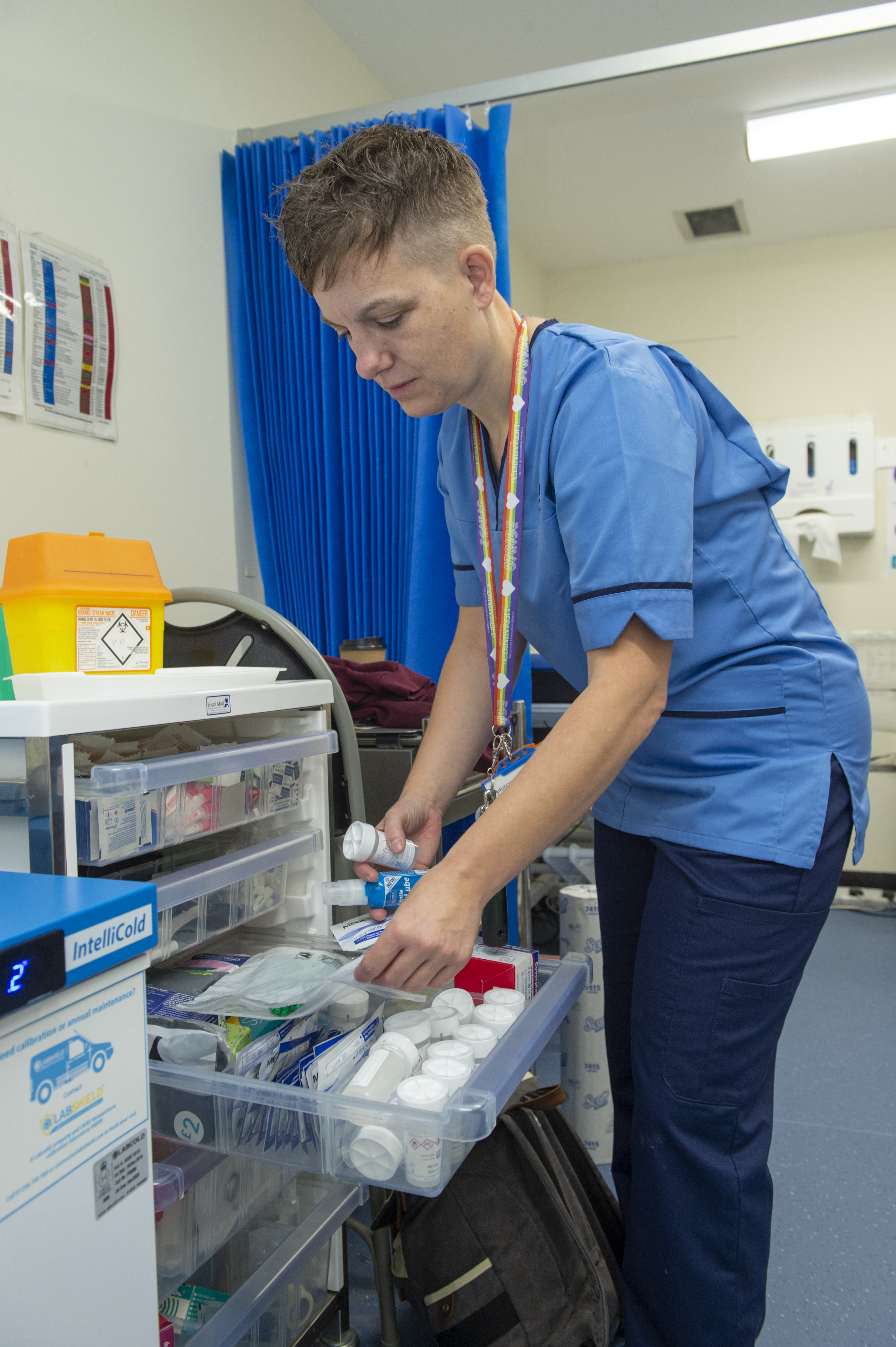 A person with short brown hair and wearing a blue nurse’s uniform of top and trousers, with a multicoloured lanyard stands by a set of perspex drawers. One drawer is open and full of bottles of pills. The person has a bottle in one hand. In the background there is an open blue curtain, cream walls and blue flooring, suggesting a clinical space.