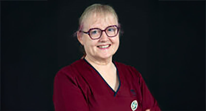 A person with light coloured hair tied back, wearing glasses and a burgundy top with dark trim, smiles at the camera against a dark background. They have their arms crossed and are wearing a pin on their top.