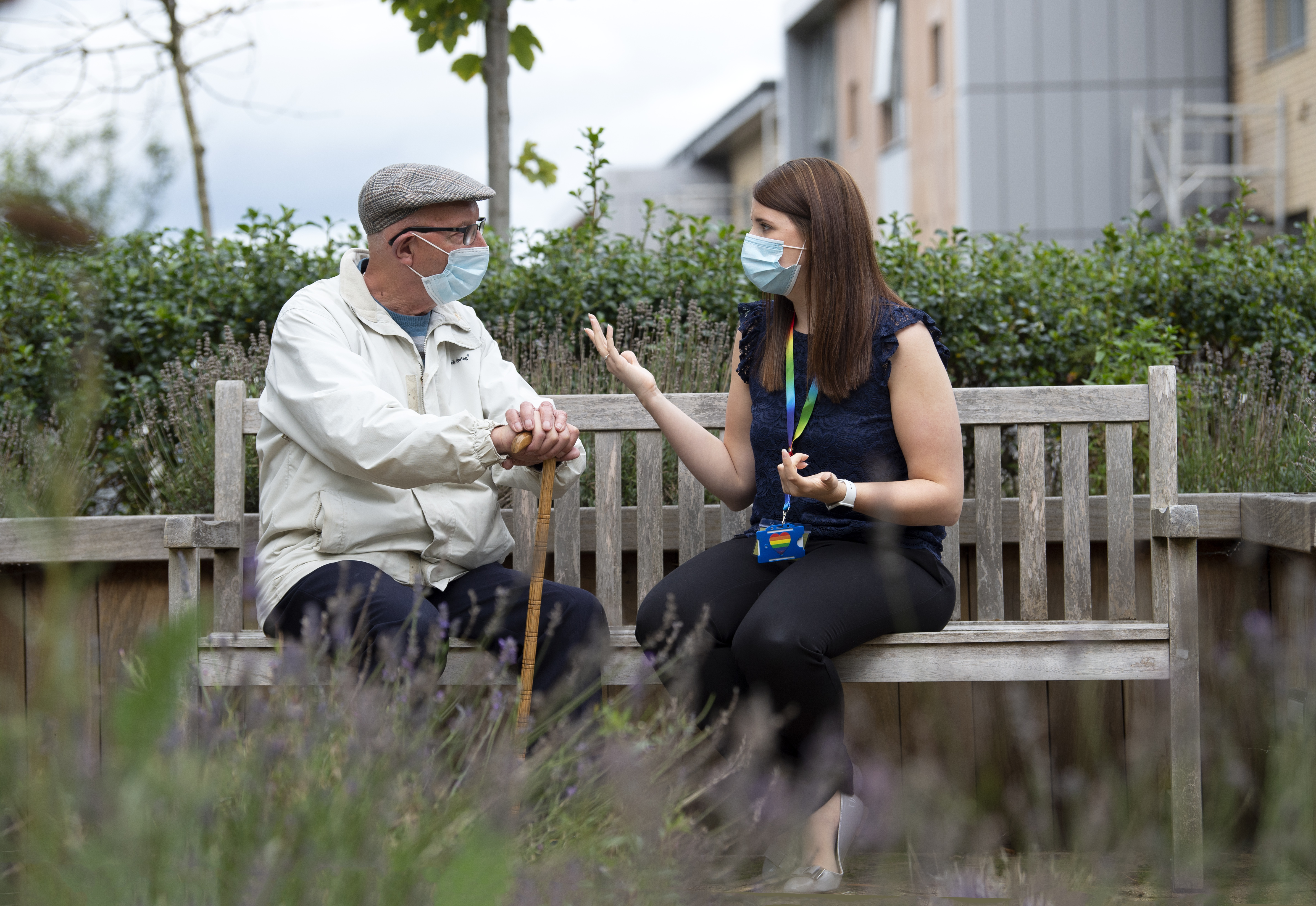 Two people sit facing each other on a wooden garden seat surrounded by trees and plants. The person on the left is wearing a grey flat cap, glasses, a cream jacket and dark trousers. They have a blue face covering on and hold a walking stick. The person on the right has long brown hair and wears a sleeveless dark blue topk, grey flat shoes with a bow on the toe and black trousers. They have a blue face covering and wear a rainbow lanyard with a blue badge attached.