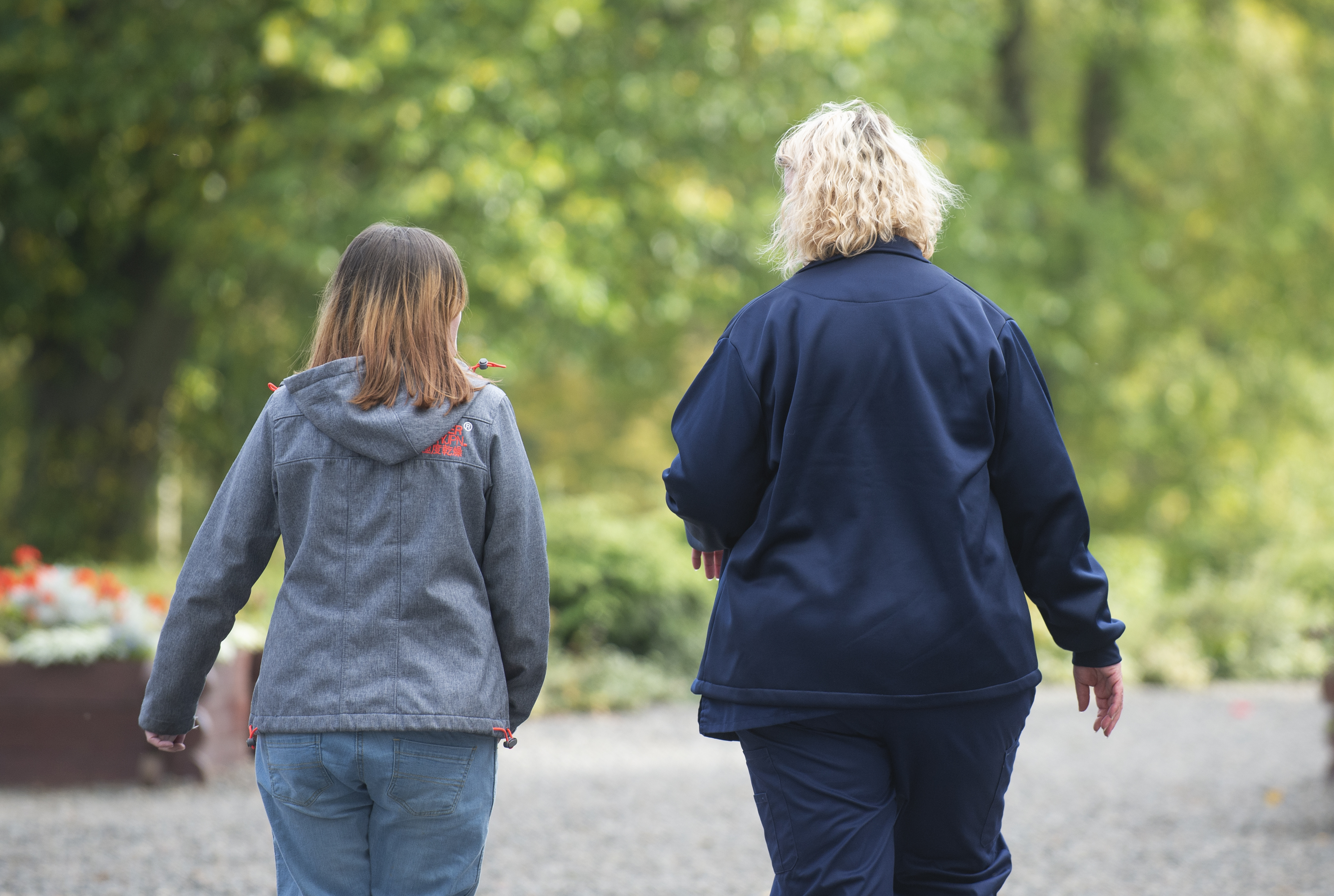 Two people walking with their backs to the camera. The person on the left has long brown hair and wears a grey hoodie and blue denim trousers. The person on the right hs curly blonde shoulder length hair. They wear a deep blue jacket and trousers. They walk on a grey path with greenery visible.